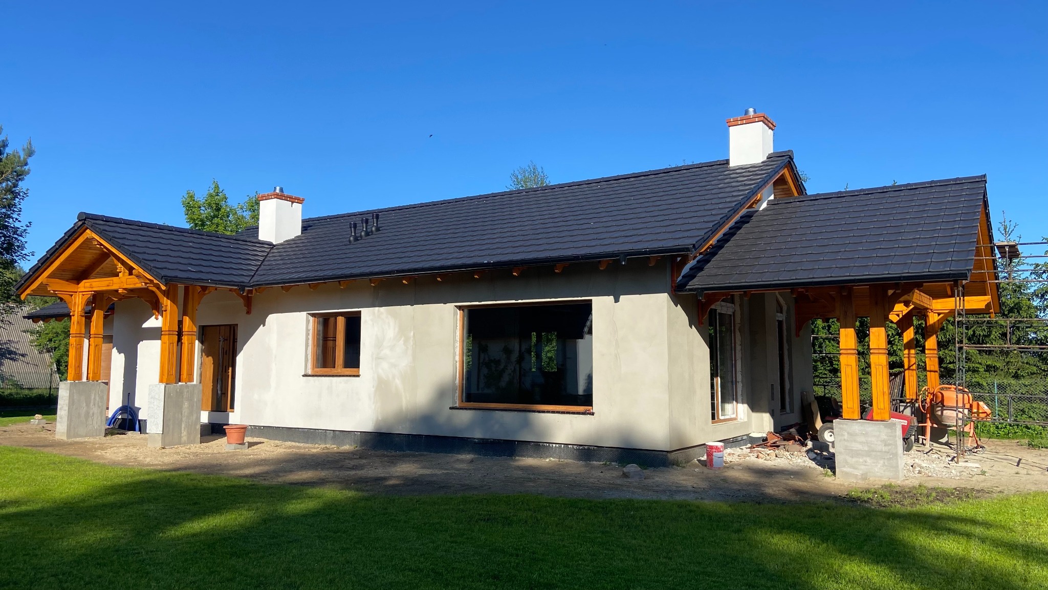 Murowany dom w trakcie wykończenia: surowe ściany, czarny dach z dachówki, drewniane kolumny werandy. Na zewnątrz betoniarka i rusztowania.