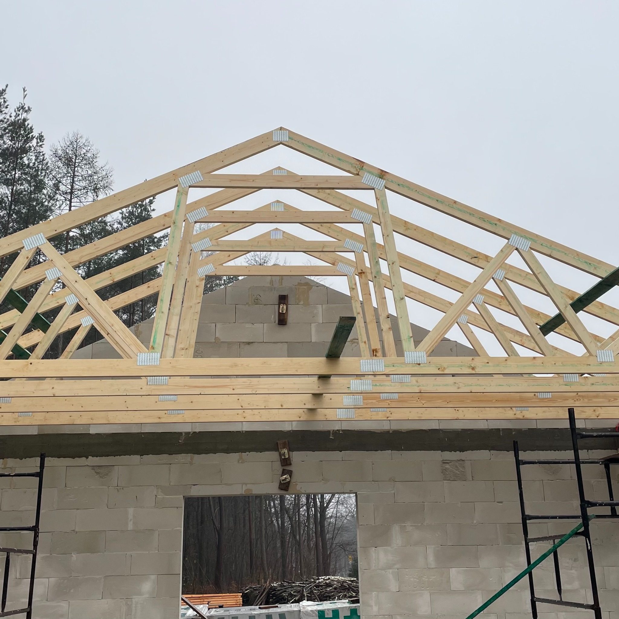 Wznoszenie konstrukcji dachu z drewnianych belek, połączonych metalowymi płytkami na budynku z białych bloczków. Widoczne rusztowania i las w tle.