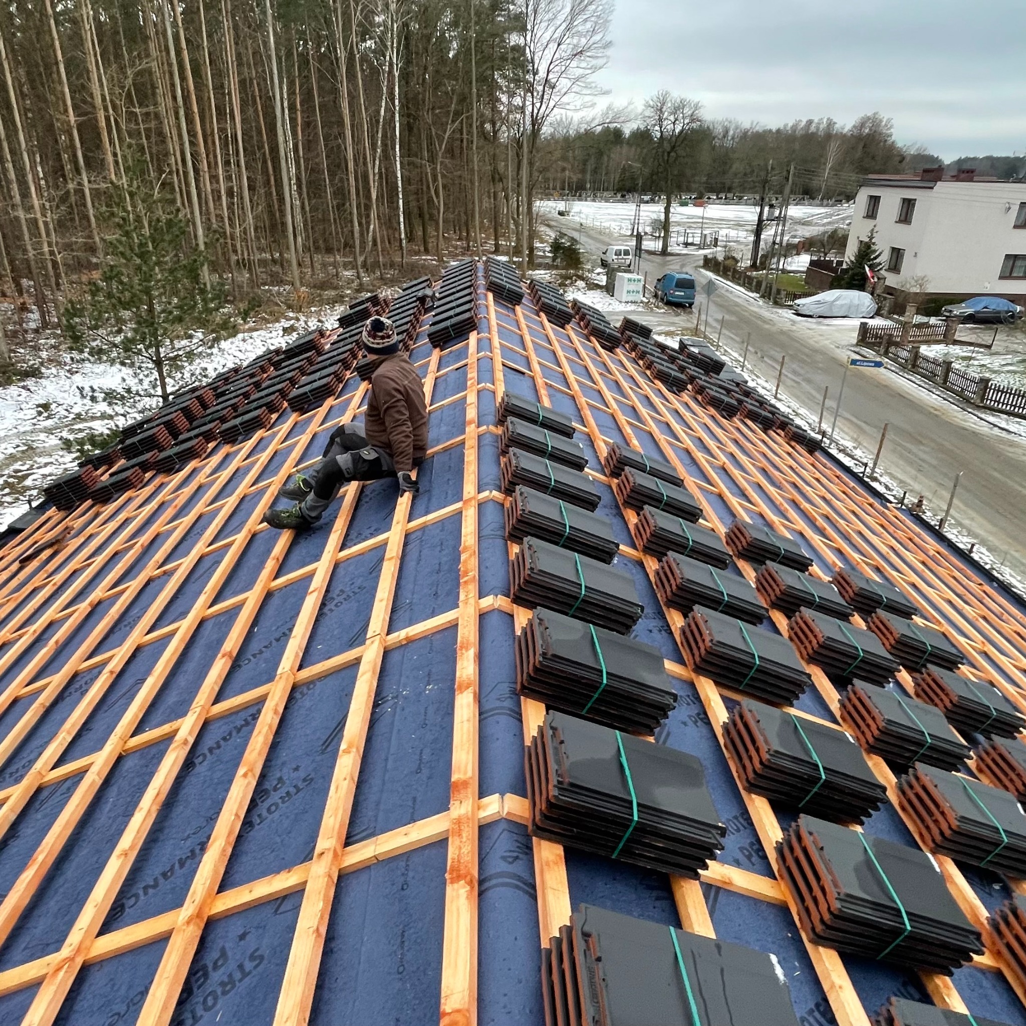 Montaż dachu z dachówki ceramicznej na drewnianej konstrukcji. Widoczny pracownik w odzieży roboczej i przygotowane paczki dachówek. Tło stanowią drzewa i zabudowa.