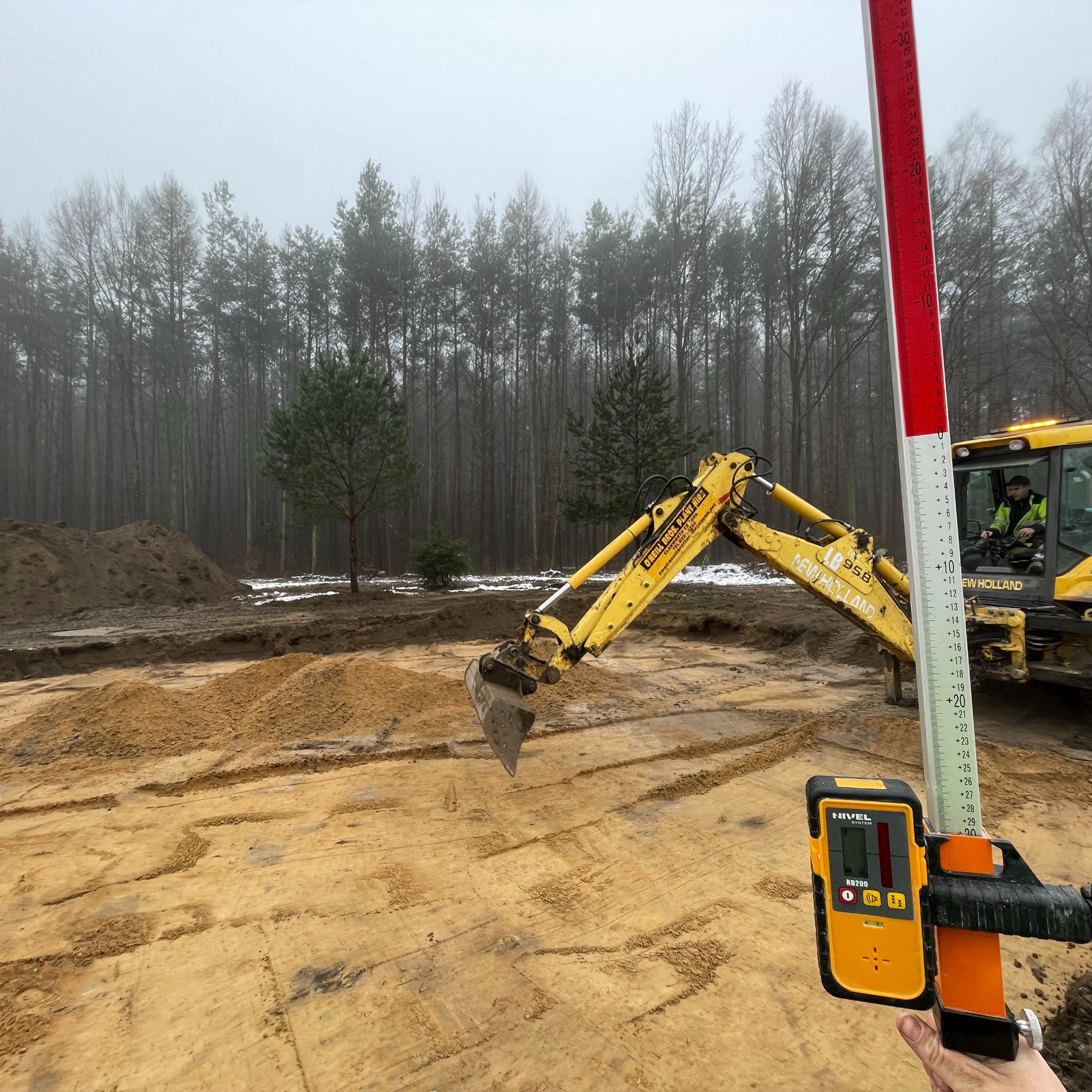 Żółta koparka New Holland w trakcie prac ziemnych na działce budowlanej, w tle las i stosy ziemi. W kadrze niwelator laserowy i łata niwelacyjna.