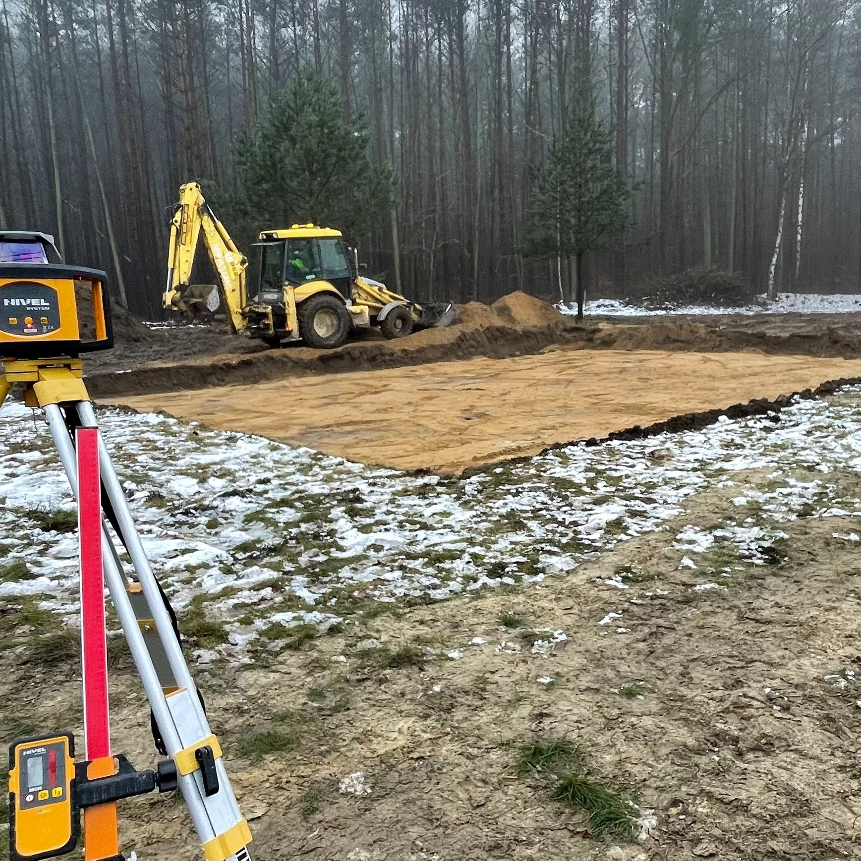 Przygotowanie terenu pod budowę domu: wykop fundamentów, żółta koparka i niwelator laserowy na trójnogu w zimowej scenerii lasu.