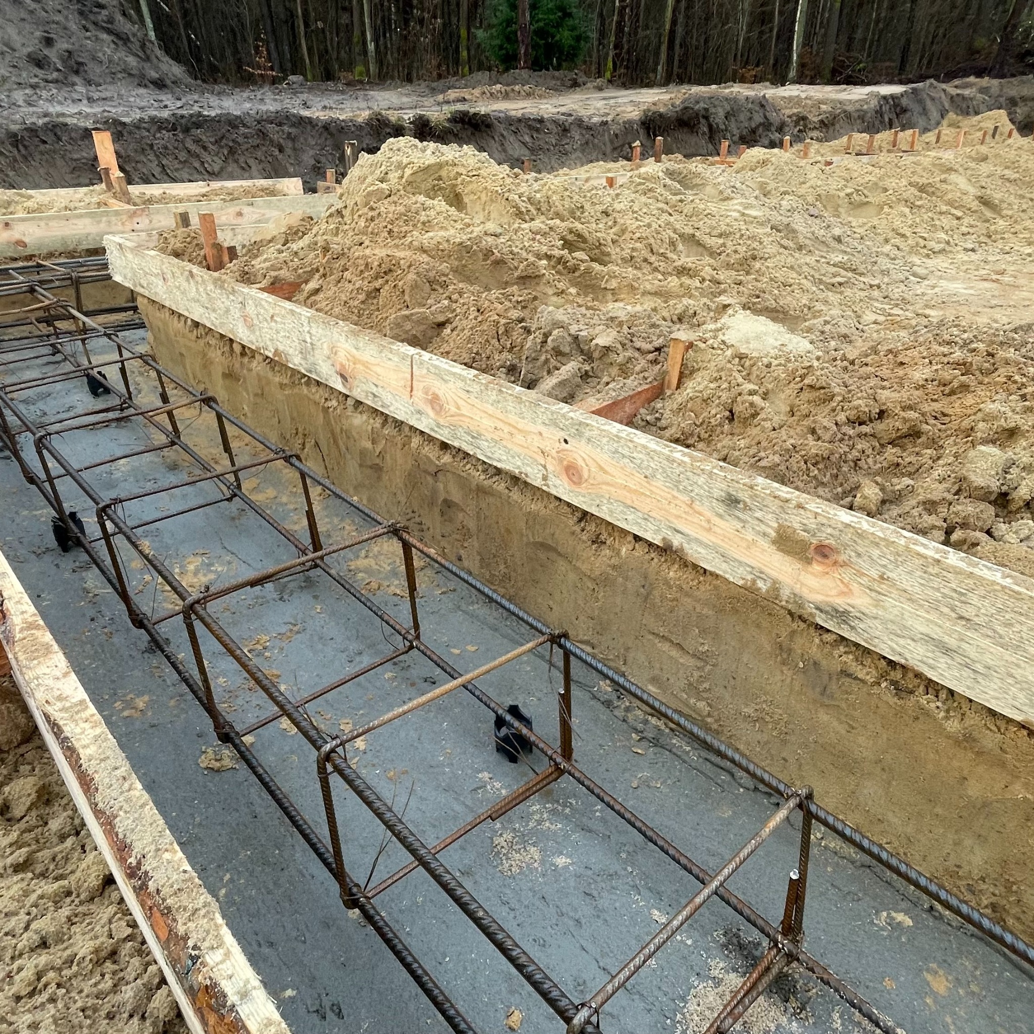 Zbrojenie fundamentów pod dom murowany, szalunek z desek, widok z góry. Przygotowanie do wylania betonu, etap budowy domu. Szare tło betonu i brązowe zbrojenie.