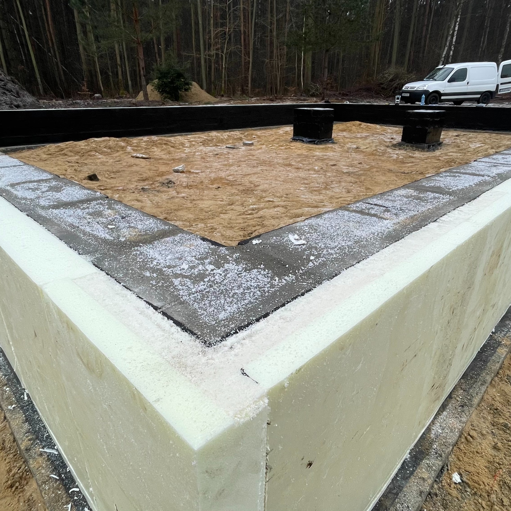 Izolowane fundamenty domu w trakcie budowy, z widoczną warstwą styropianu i piaskiem wewnątrz. W tle las i biały samochód dostawczy.
