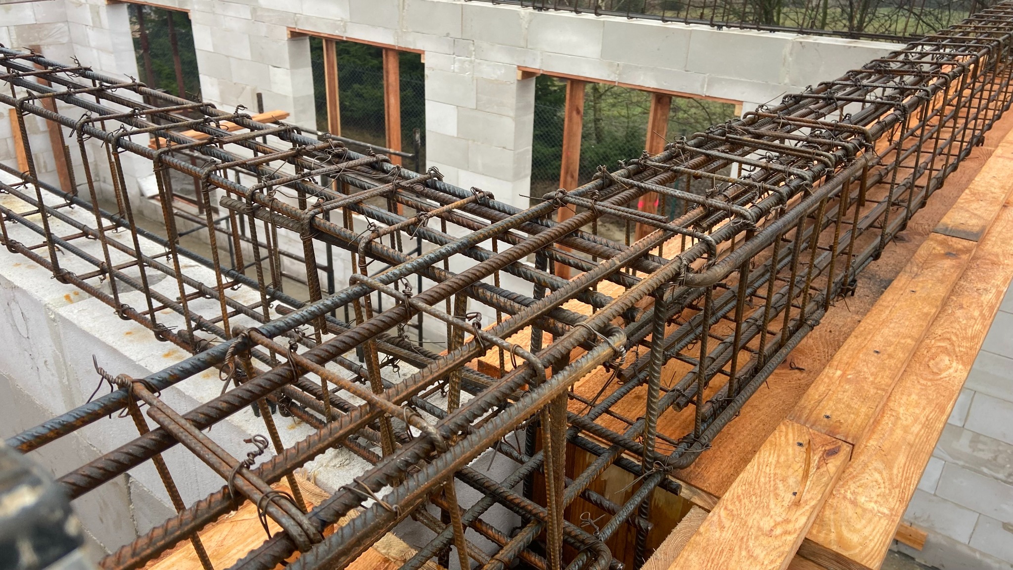 Zbrojenie belki stropowej na deskowaniu, widoczne bloczki betonowe i elementy konstrukcji domu w budowie. Detal z bliska, surowy stan.