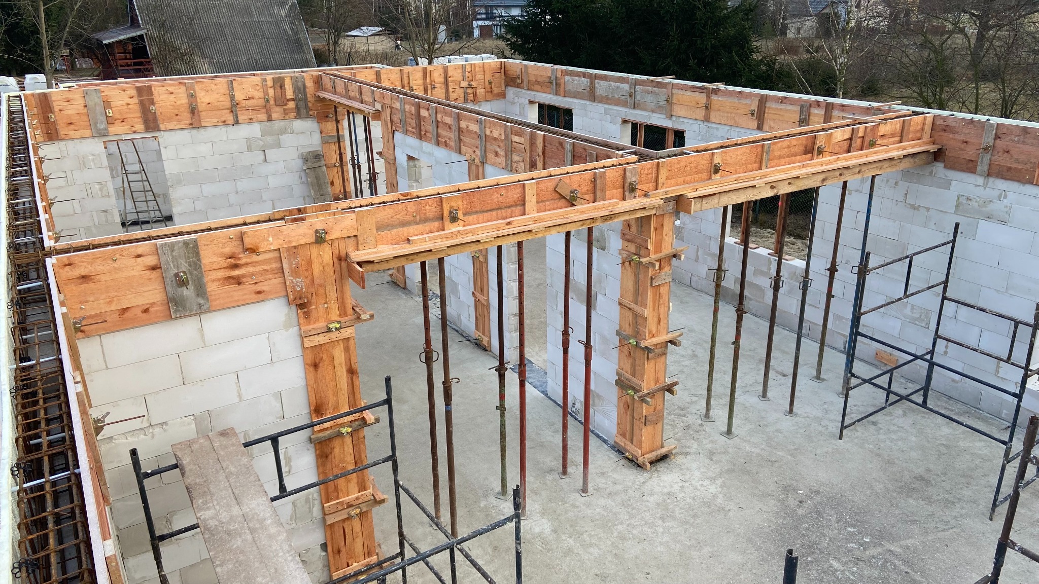 Wznoszenie ścian domu z bloczków, drewniane szalunki podtrzymują konstrukcję. Widoczne rusztowania i stalowe podpory. Stan surowy otwarty, w trakcie budowy.