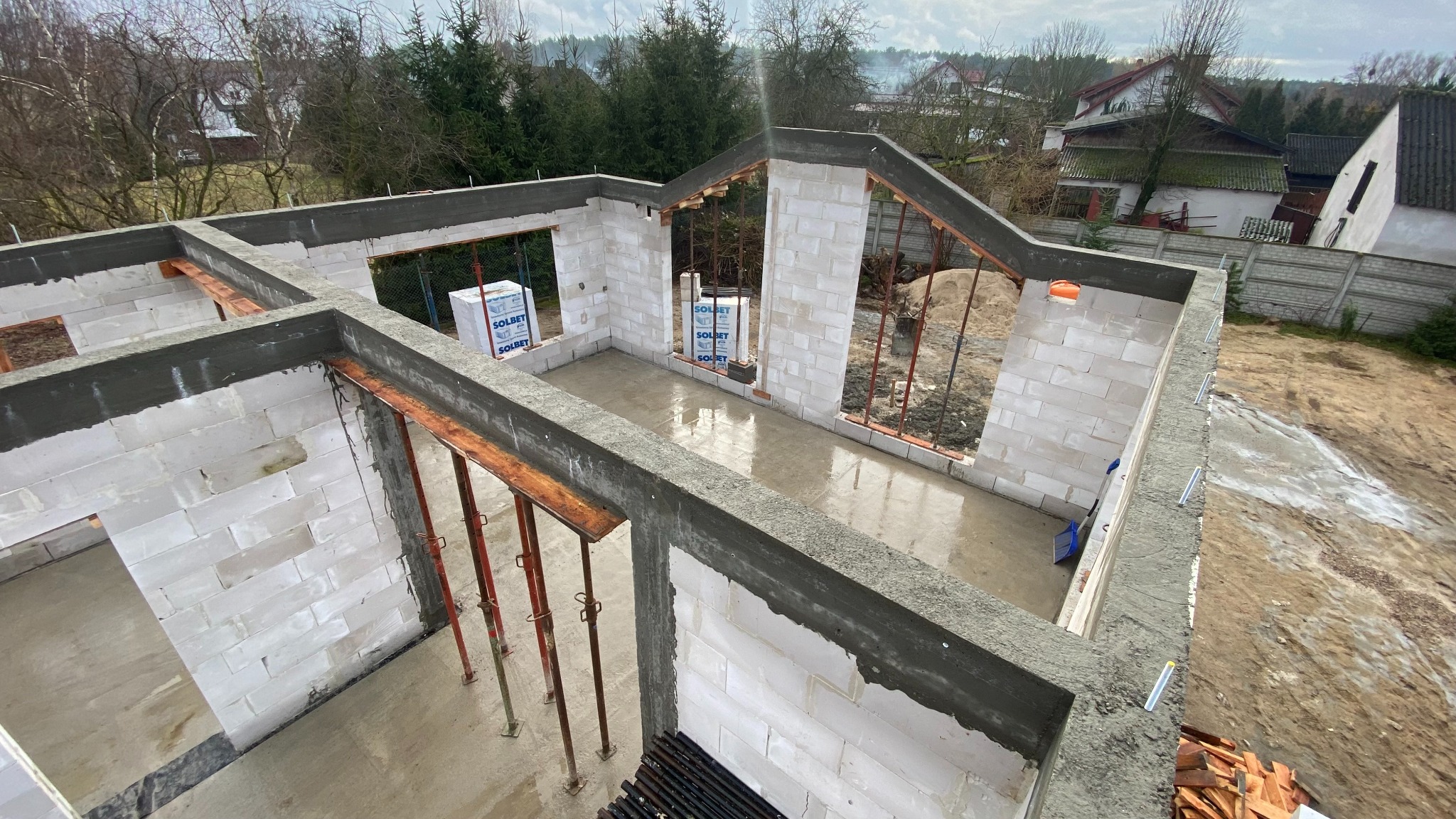 Wysoki kąt na fundamenty domu z białego bloczka Solbet, widoczne podpory i mokra wylewka. W tle drzewa i zabudowa jednorodzinna.