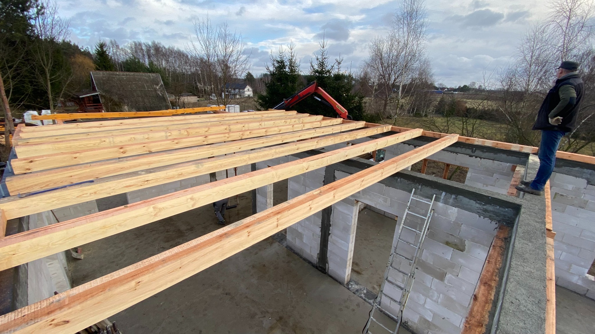 Budowa domu z pustaków: widok z góry na drewniane belki stropowe, częściowo wzniesione ściany i pracownika na krawędzi. W tle las i krajobraz.