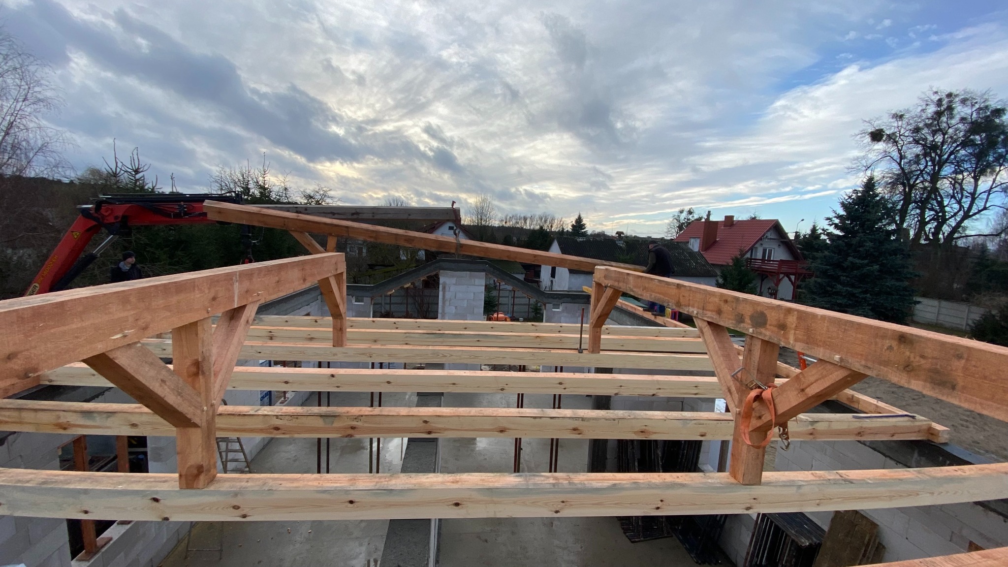 Budowa domu murowanego w Grudziądzu: widok z góry na konstrukcję dachu z drewnianych belek, na tle nieba i zabudowań. Prace w toku, dźwig w tle.