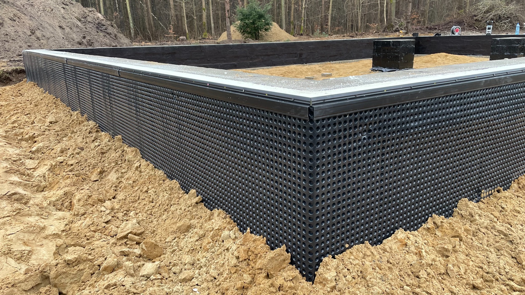 Fundament domu z czarną folią kubełkową i piaskiem. Widoczne elementy konstrukcyjne. Na tle las.