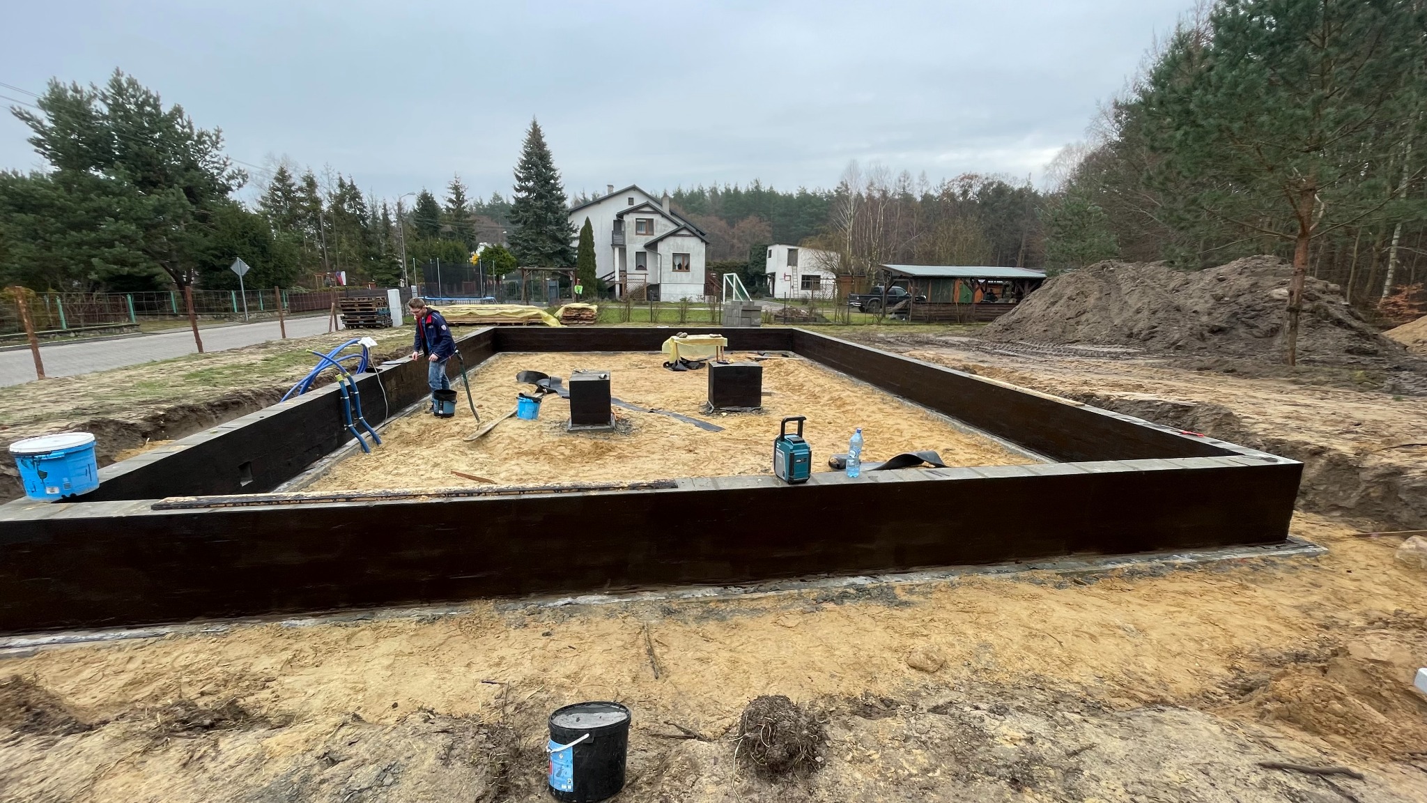 Przygotowanie fundamentów pod dom murowany: zalane ławy fundamentowe, izolacja pionowa, piasek zasypany wewnątrz. W tle dom jednorodzinny i drzewa.