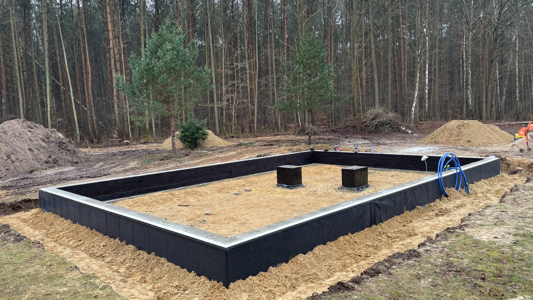 Fundament domu murowanego z izolacją pionową na tle lasu. Widoczne dwa czarne słupki, piasek wewnątrz i pryzmy ziemi dookoła.