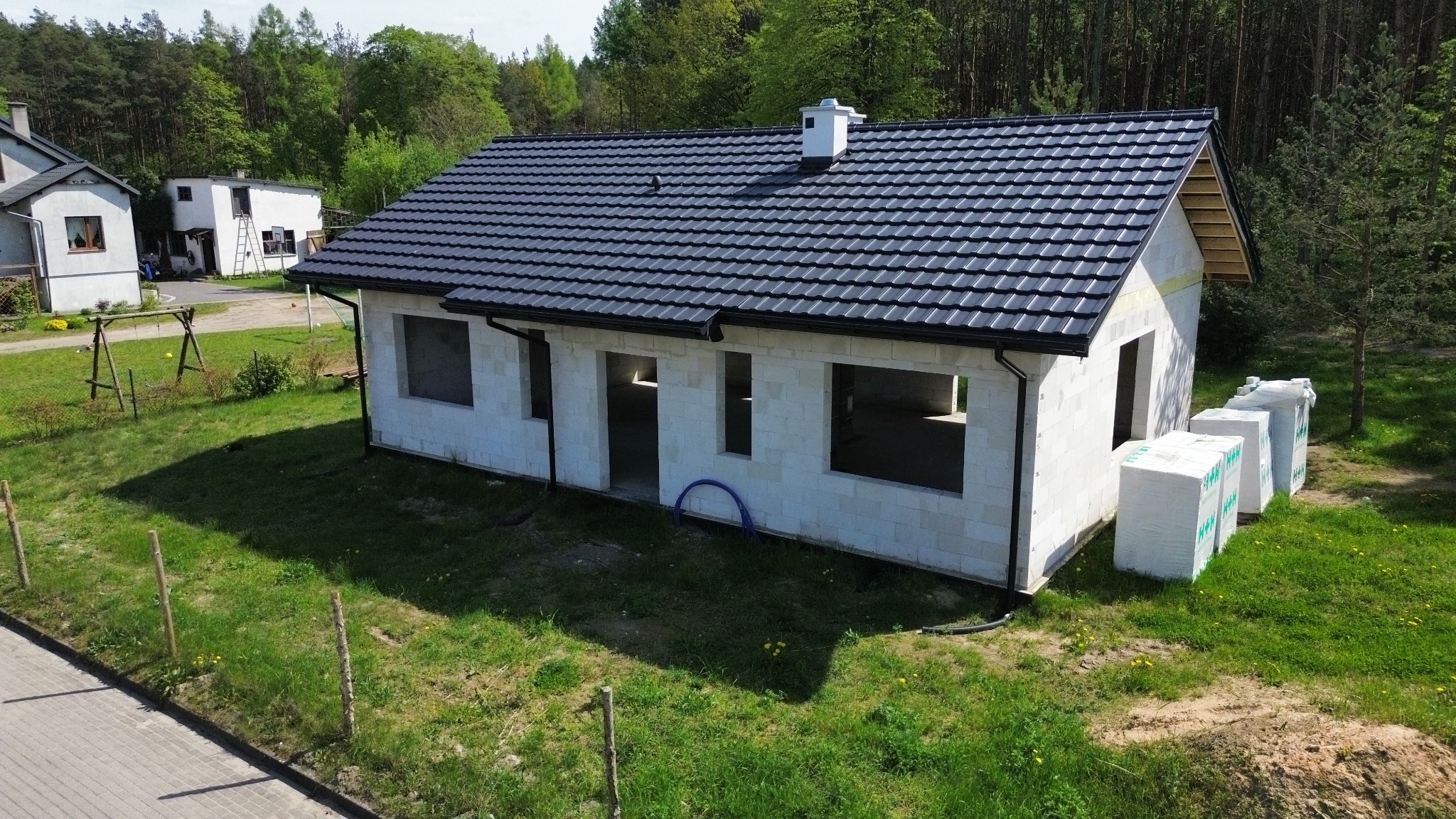 Stan surowy otwarty domu jednorodzinnego z ciemną dachówką, białe bloczki, widoczne materiały budowlane obok, zielone otoczenie i las w tle.