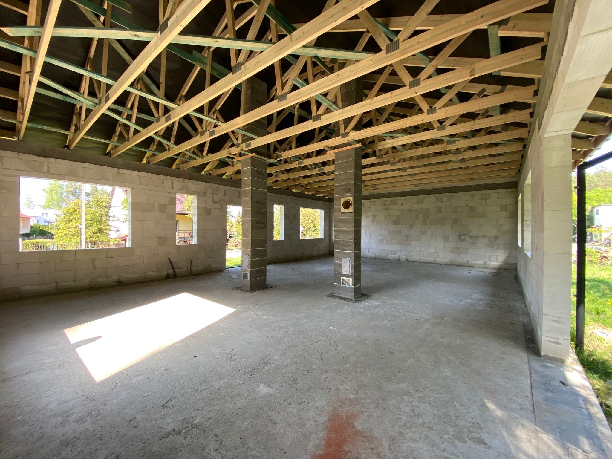 Surowy stan domu murowanego: ściany z bloczków, betonowa podłoga, drewniana konstrukcja dachu z zielonymi elementami. Widok na zewnątrz przez otwory okienne.