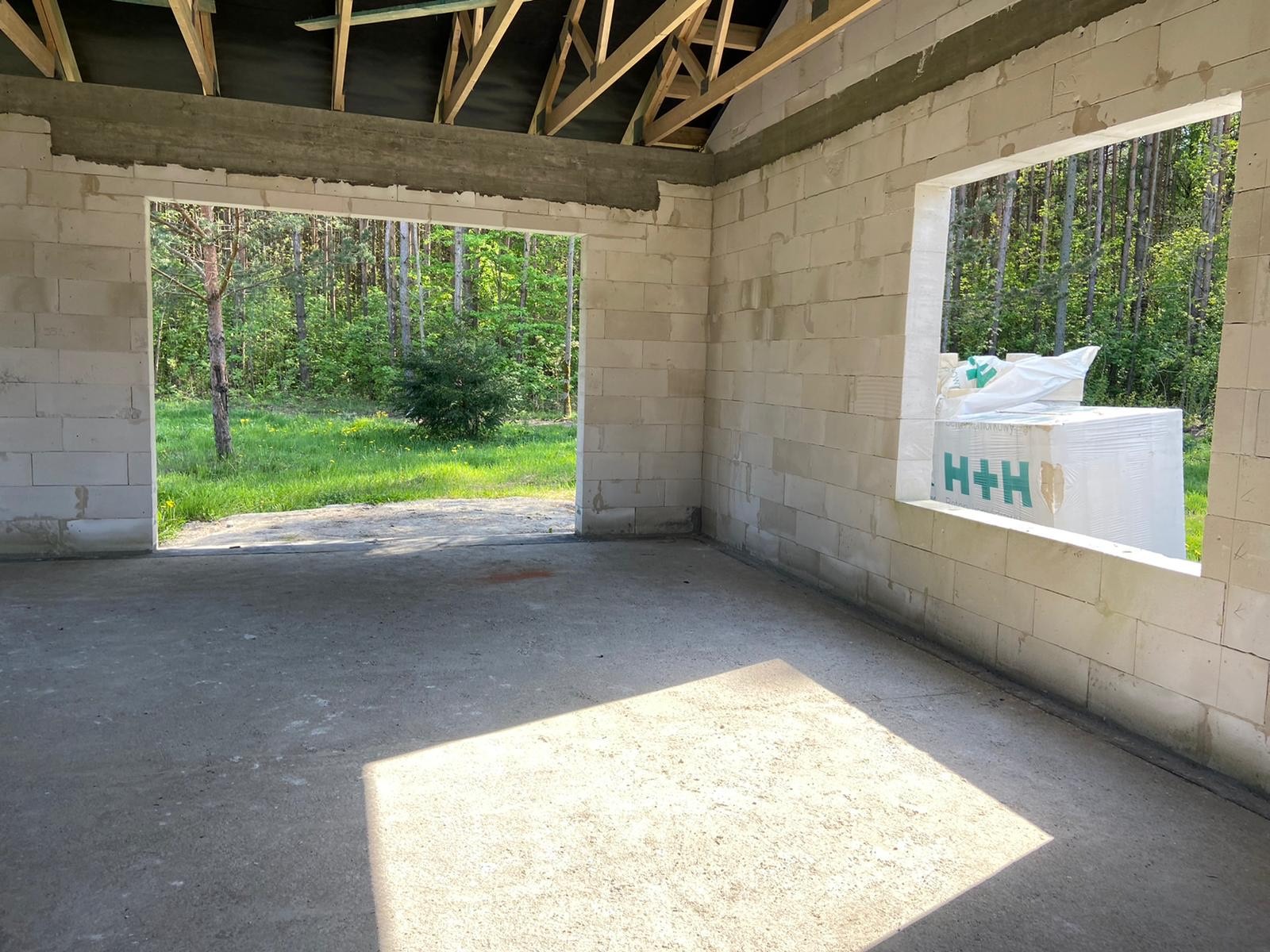 Wnętrze surowego domu z bloczków H+H, widok na las przez otwór okienny i drzwiowy. Elementy więźby dachowej widoczne. Podłoga betonowa, wpadające światło słoneczne.