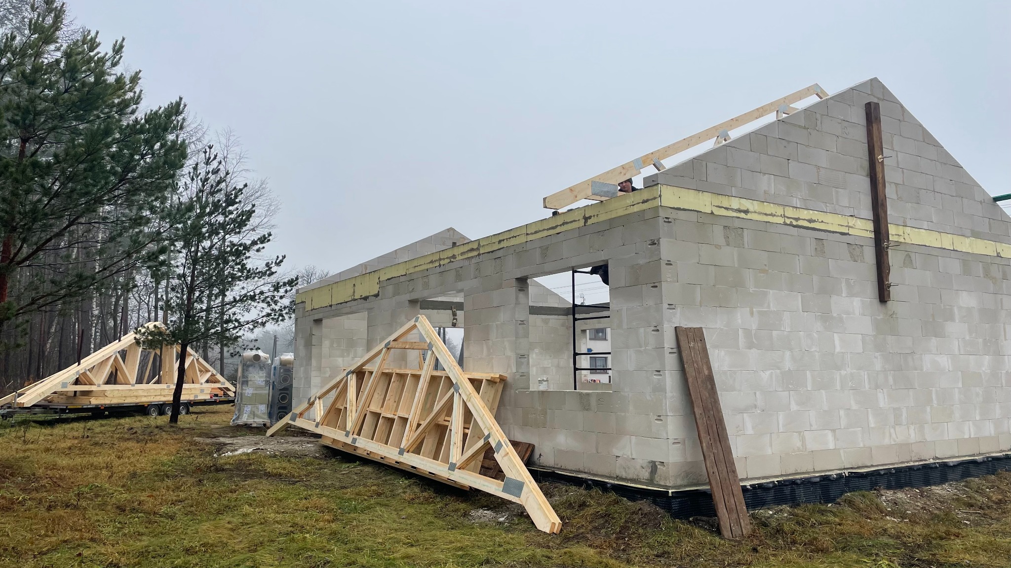 Budowa domu z bloczków: ściany wzniesione, przygotowane krokwie dachowe, widoczna izolacja. Etap montażu więźby dachowej na tle pochmurnego nieba.