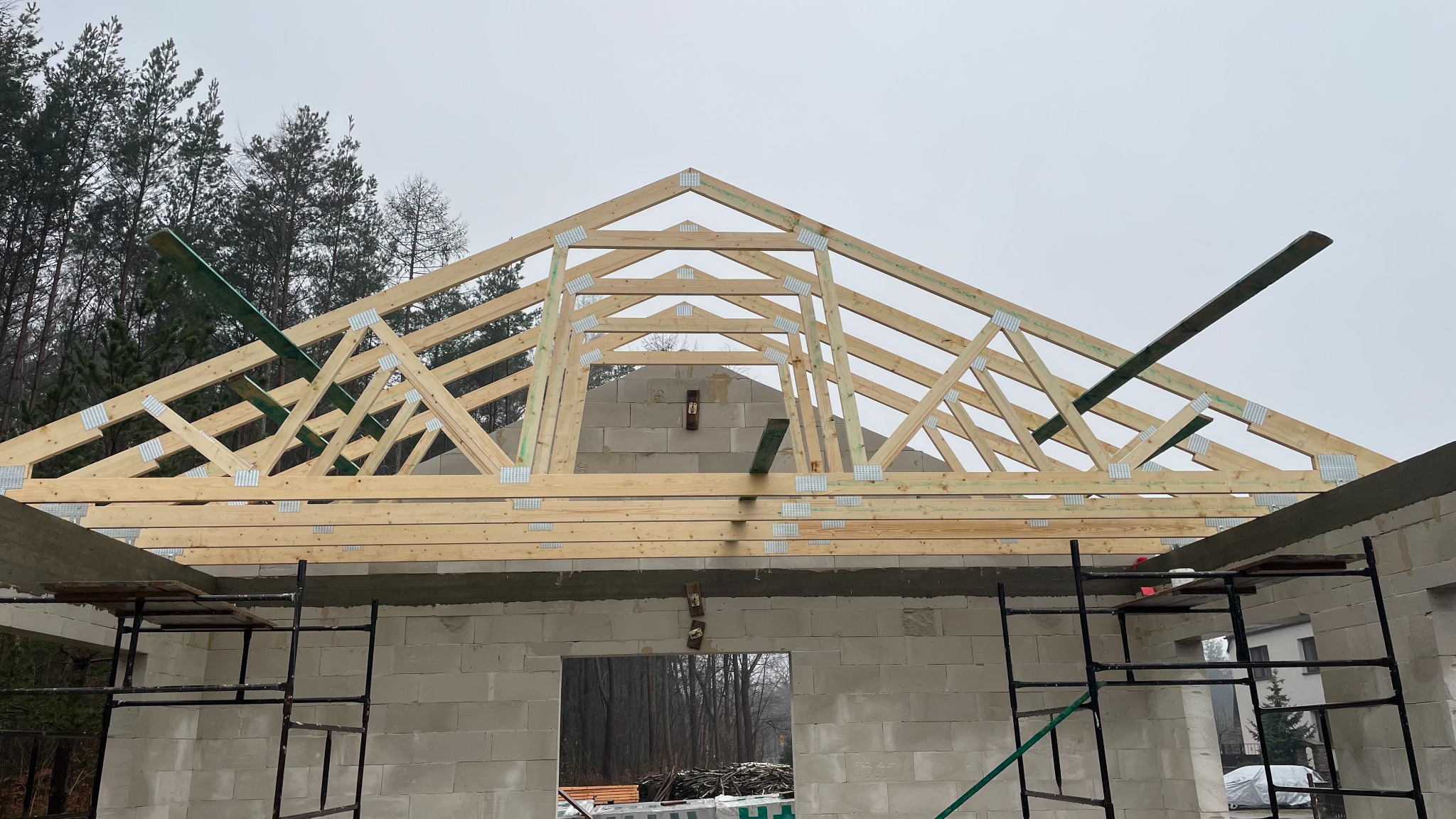 Drewniana konstrukcja dachu z elementami metalowymi na murowanym domu w trakcie budowy. Widoczne rusztowania i drzewa w tle.