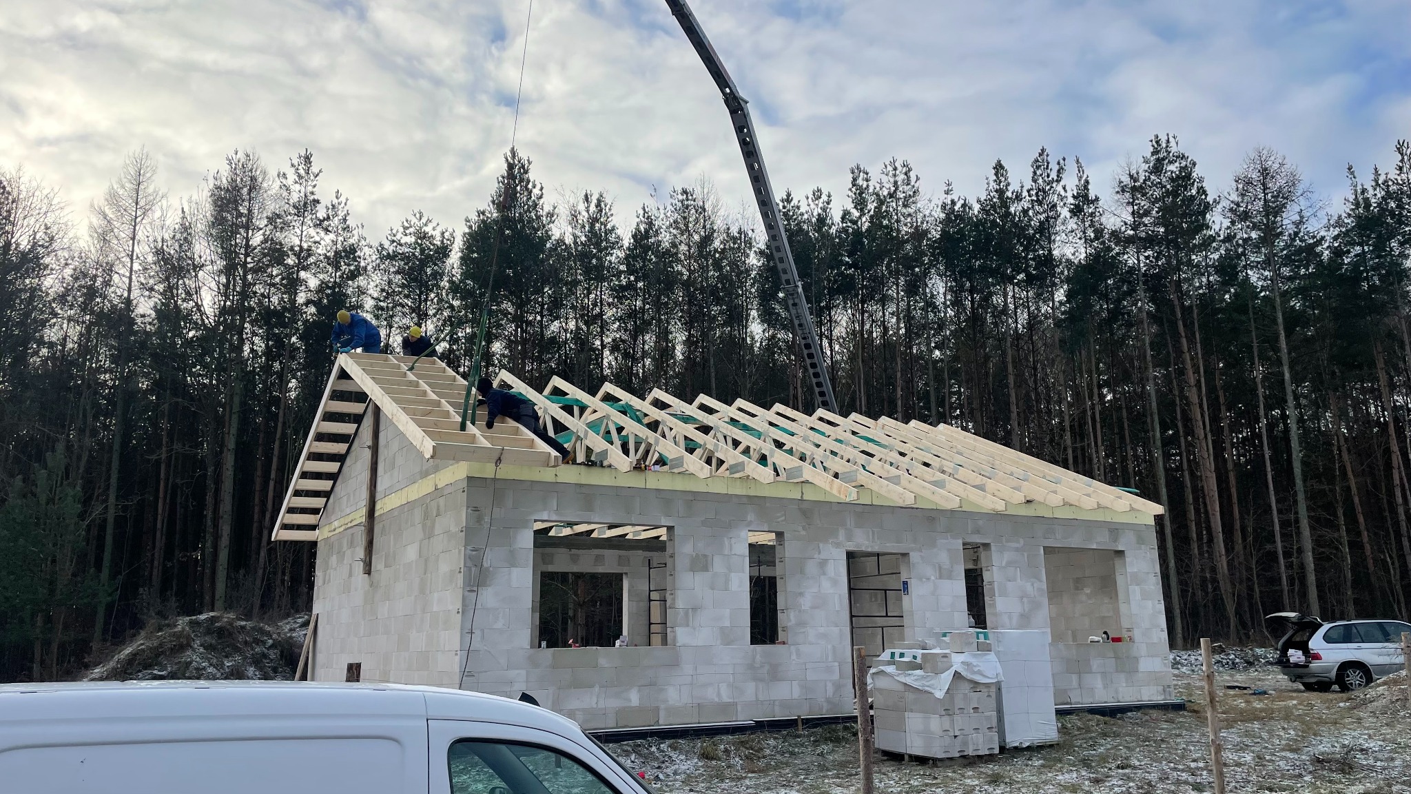 Budowa domu z bloczków: ekipa montuje drewnianą konstrukcję dachu, z tyłu las iglasty, na pierwszym planie częściowo widoczny samochód dostawczy.