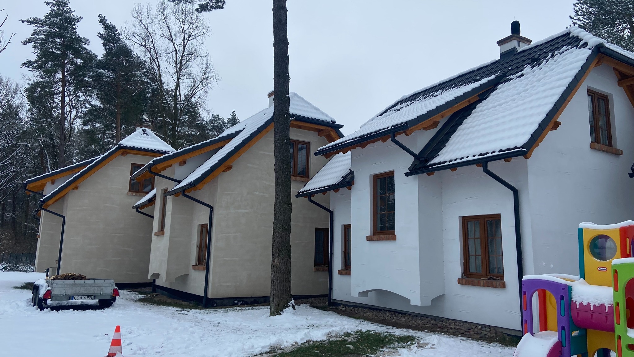 Zimowy krajobraz z trzema domami w różnym stadium wykończenia. Jeden dom jest biały, drugi w trakcie wykończenia, a trzeci w kolorze beżowym. Na dachu leży śnieg.