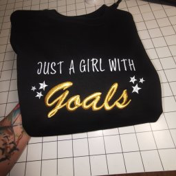 MARTYNA SZWED - Czarna bluza z haftowanym napisem 'JUST A GIRL WITH Goals' w kolorze białym i złotym, leżąca na białym stole z czarną kratką. Widoczny tatuaż na ramieniu.