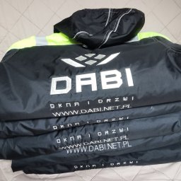 MARTYNA SZWED - Stos kurtek roboczych z logo firmy DABI, w kolorze czarnym z neonowymi wstawkami, leżących na pikowanej narzucie. Widoczne nadrukowane dane kontaktowe.