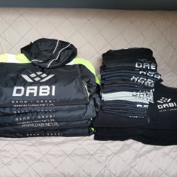 MARTYNA SZWED - Stos odzieży roboczej z nadrukowanym logo firmy DABI. Czarna kurtka z elementami neonowymi i czarne bluzy leżą na jasnym tle.