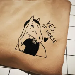 Beżowa bluza z haftem przedstawiającym konia w koszuli i napisem 'Yes of Horse' leży na białej, kafelkowej powierzchni.