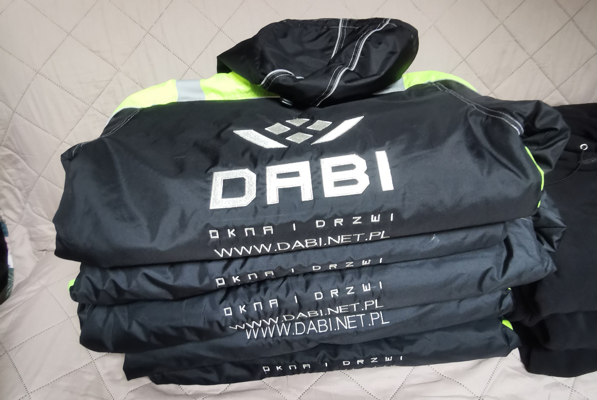 Stos kurtek roboczych z logo firmy DABI, w kolorze czarnym z neonowymi wstawkami, leżących na pikowanej narzucie. Widoczne nadrukowane dane kontaktowe.