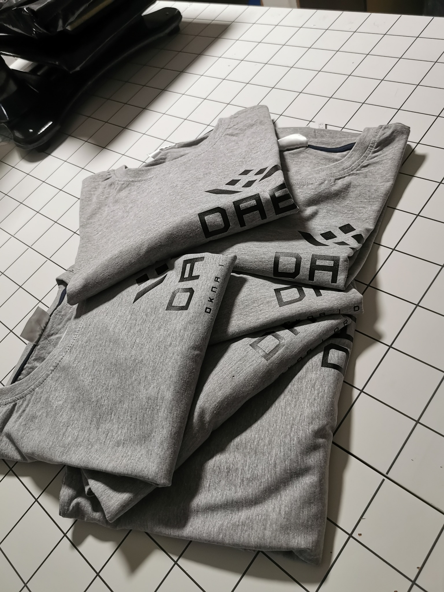 Stos szarych koszulek z czarnym nadrukiem logo na rękawie, ułożonych na białym stole z kratką. Widoczne detale materiału i precyzja nadruku.