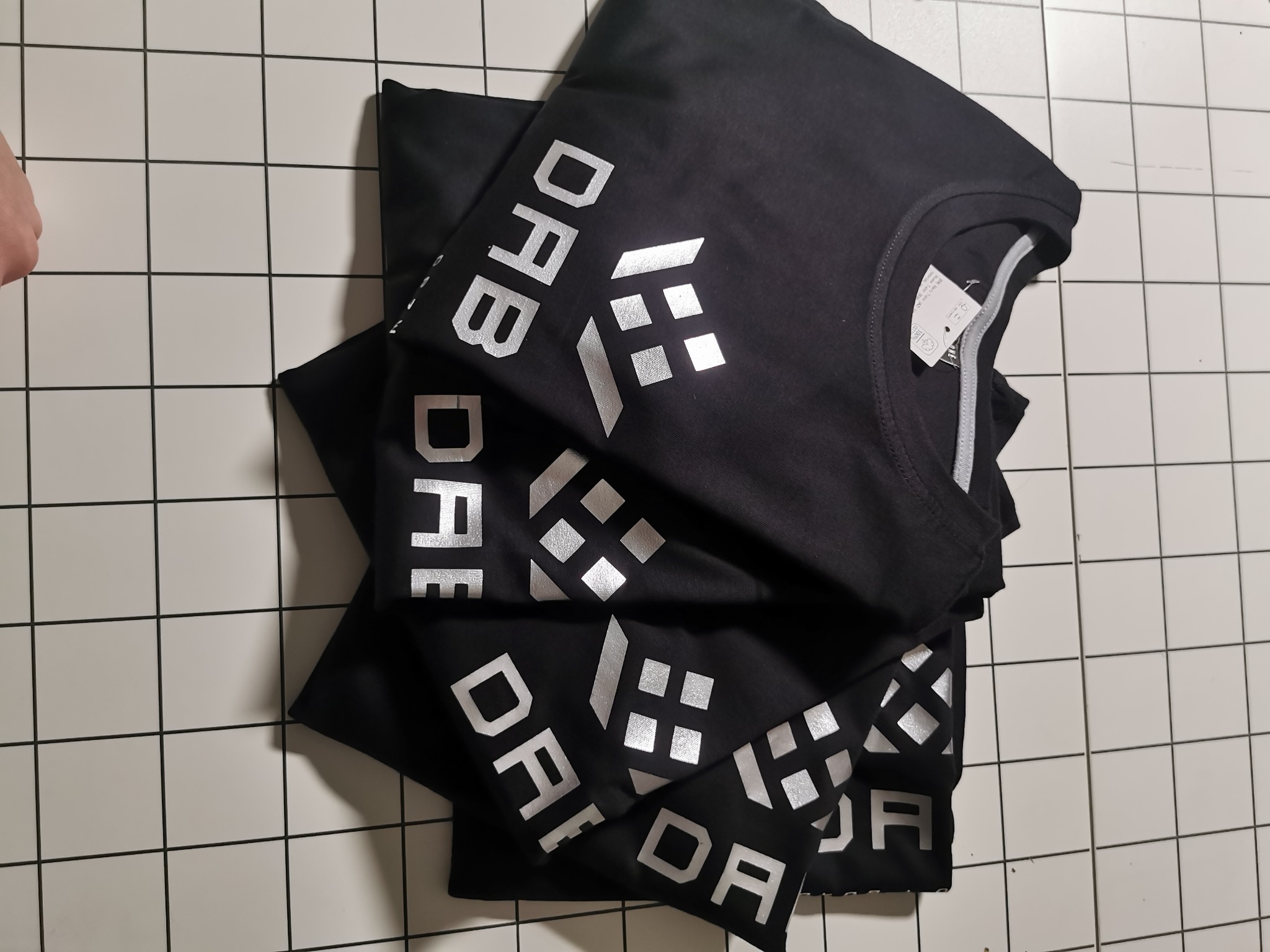 Stos czarnych koszulek z srebrnym nadrukiem 'DAFB' na kafelkowanym tle. Widoczny metka na jednej z koszulek. Ujęcie z góry, lekko z ukosa.