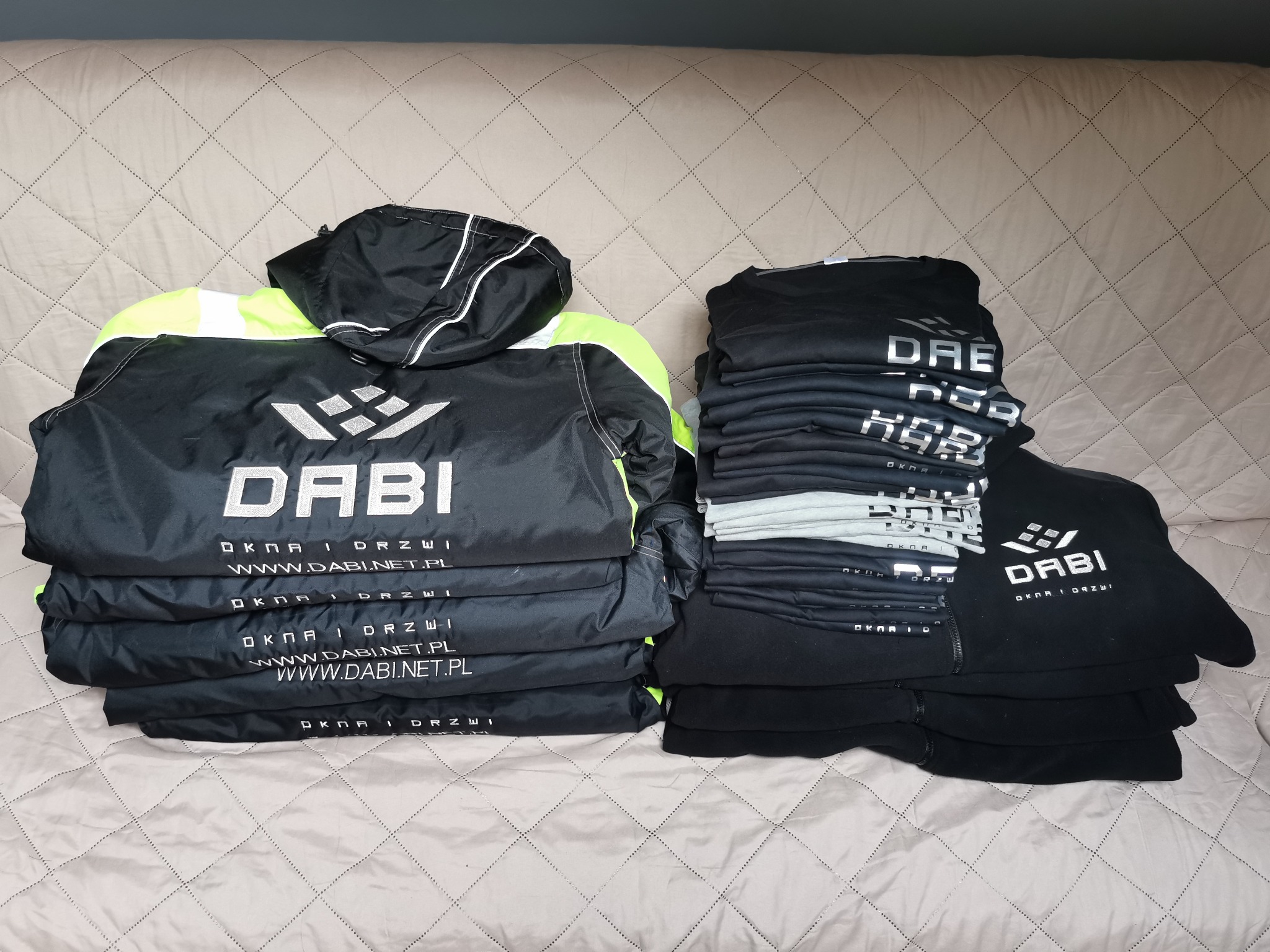 Stos odzieży roboczej z nadrukowanym logo firmy DABI. Czarna kurtka z elementami neonowymi i czarne bluzy leżą na jasnym tle.