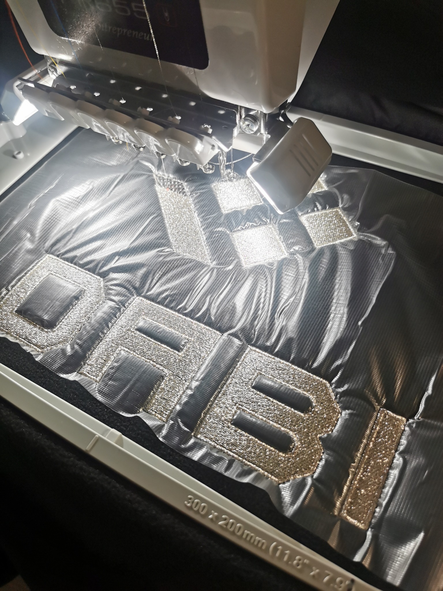 Proces haftowania srebrzystego napisu 'DABI' na czarnym materiale, widoczny mechanizm maszyny hafciarskiej i błyszczące nici w trakcie pracy.