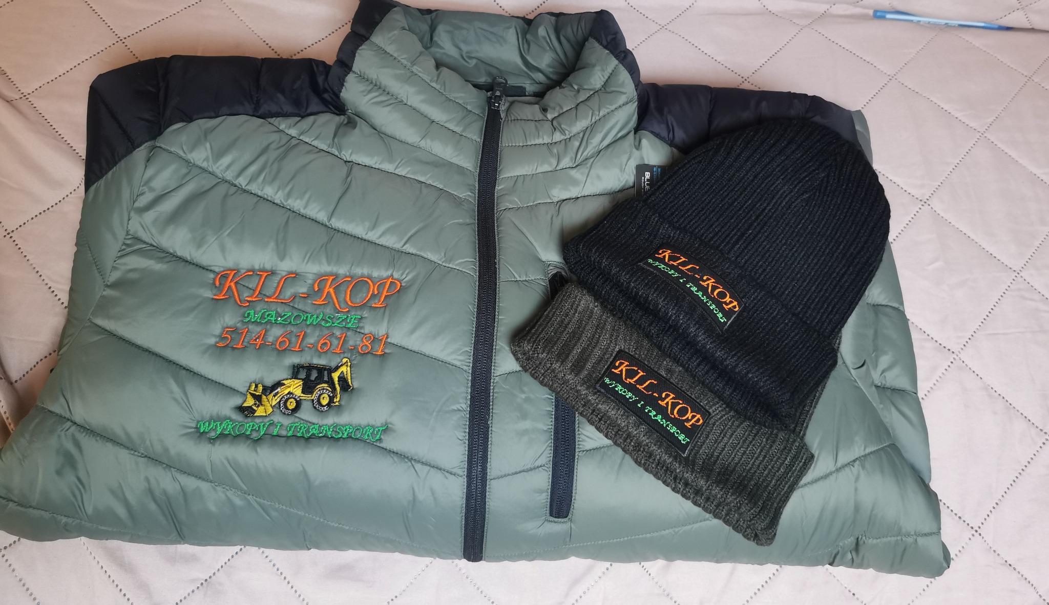 Oliwkowa kurtka i czapka z haftowanym logo firmy budowlanej 'KIL-KOP' z Mazowsza, z numerem telefonu i grafiką koparki. Prezentacja odzieży roboczej z haftem.
