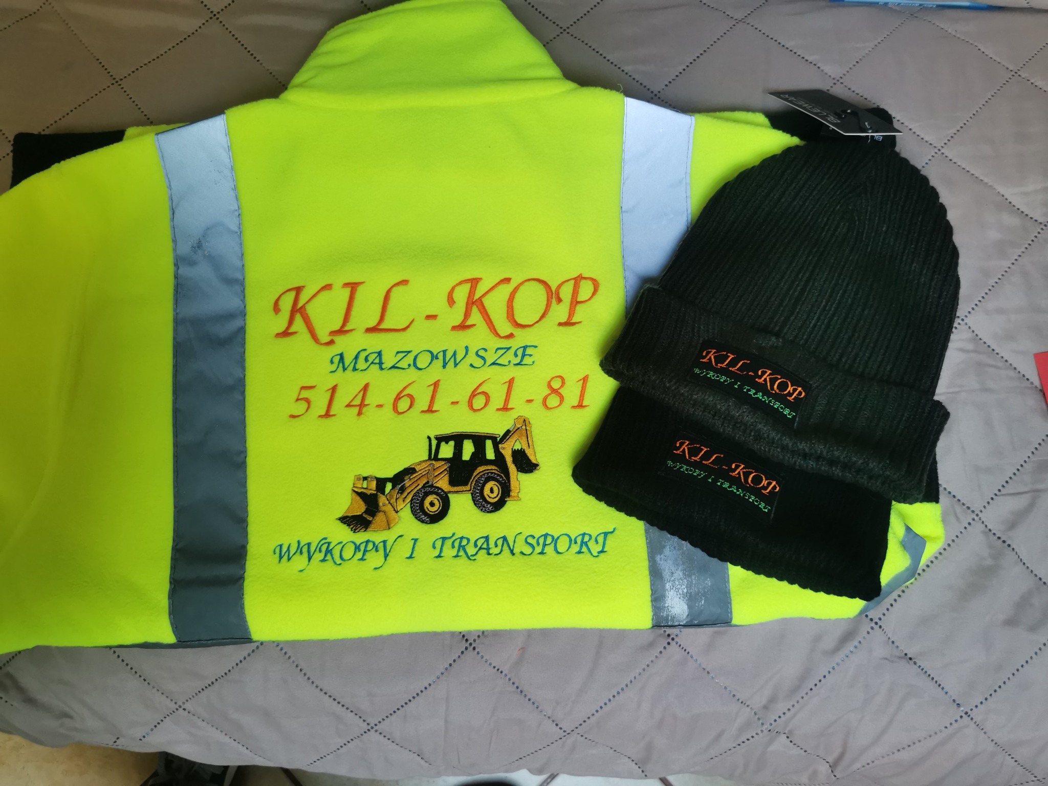 Żółta bluza robocza z haftowanym logo 'KIL-KOP' i numerem telefonu, obok czapka z tym samym haftem. Odzież z logo firmy na szarym tle.