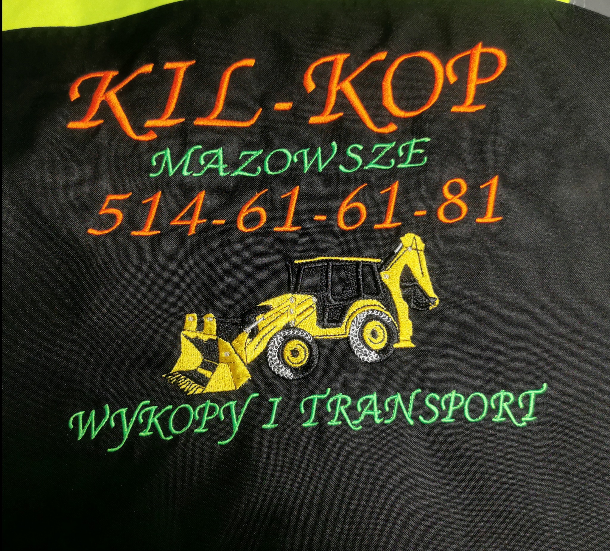 Czarna tkanina z kolorowym haftem: logo firmy KIL-KOP, Mazowsze, numer telefonu i grafika koparki. Wykopy i transport. Detal haftu na odzieży roboczej.