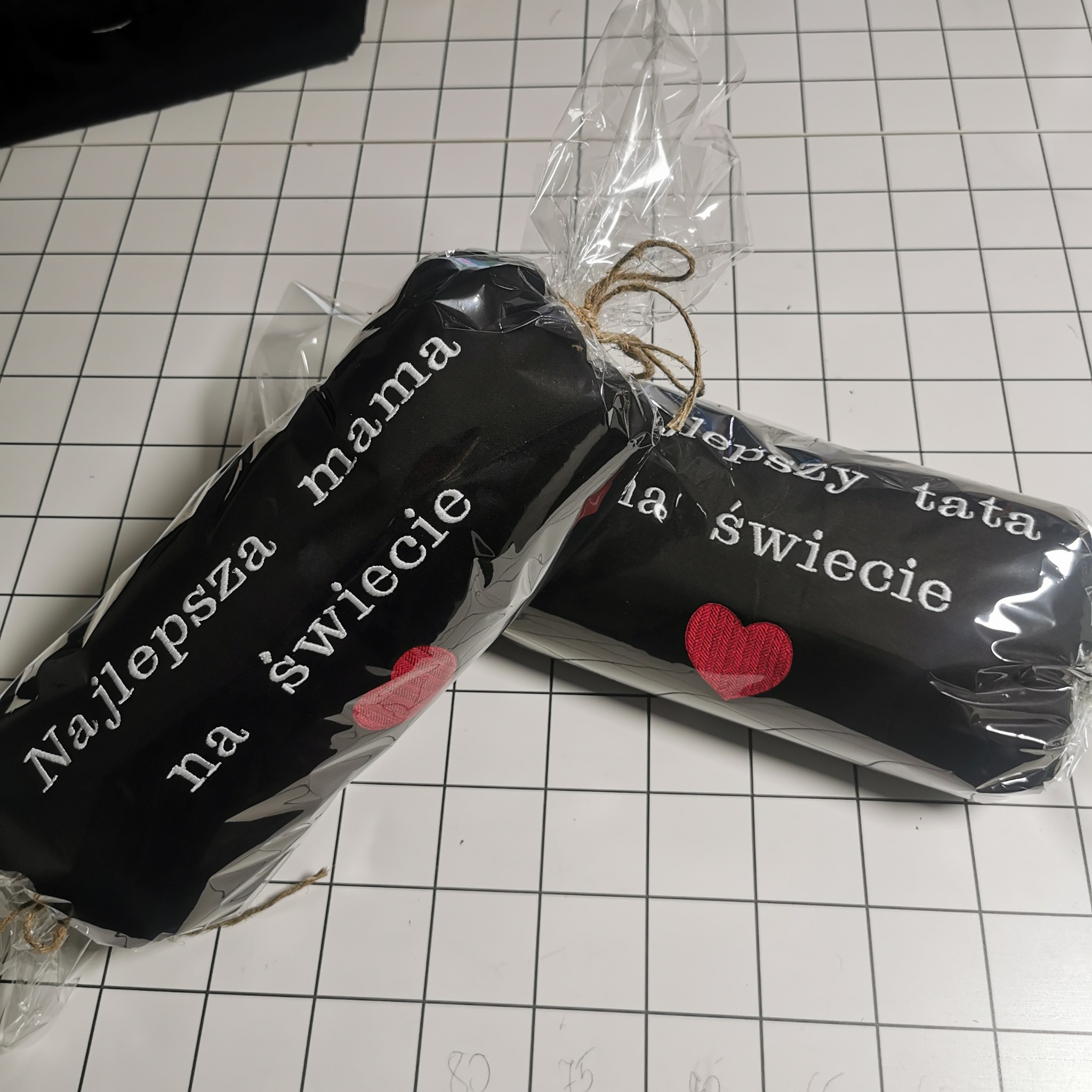 Dwa czarne ręczniki z haftowanymi napisami 'Najlepsza mama na świecie' i 'Najlepszy tata na świecie', ozdobione czerwonymi sercami, zapakowane w folię i związane sznurkiem.