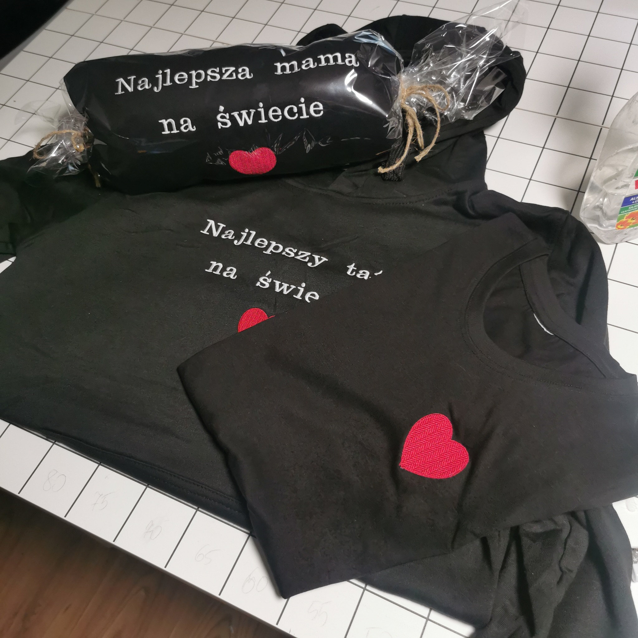 Czarne ubrania z haftowanymi napisami i sercami na tle białej kratki. Bluza z kapturem i koszulka z napisem 'Najlepszy tata na świecie'. Poduszka z napisem 'Najlepsza mama'.
