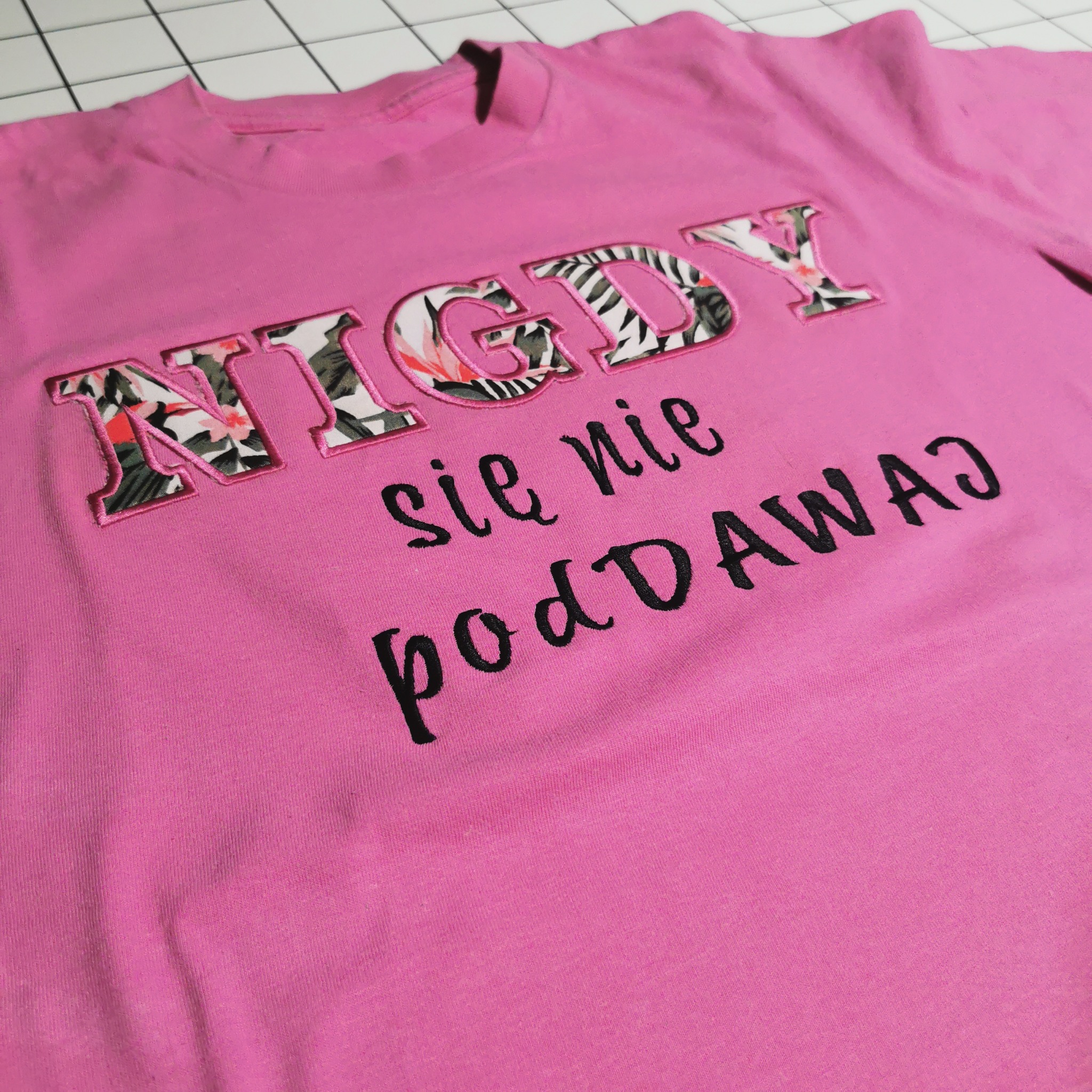 Różowa koszulka z haftowanym napisem 'NIGDY się nie podDAWAJ', litery wypełnione wzorem kwiatowym. Prezentacja nadruku na odzieży.