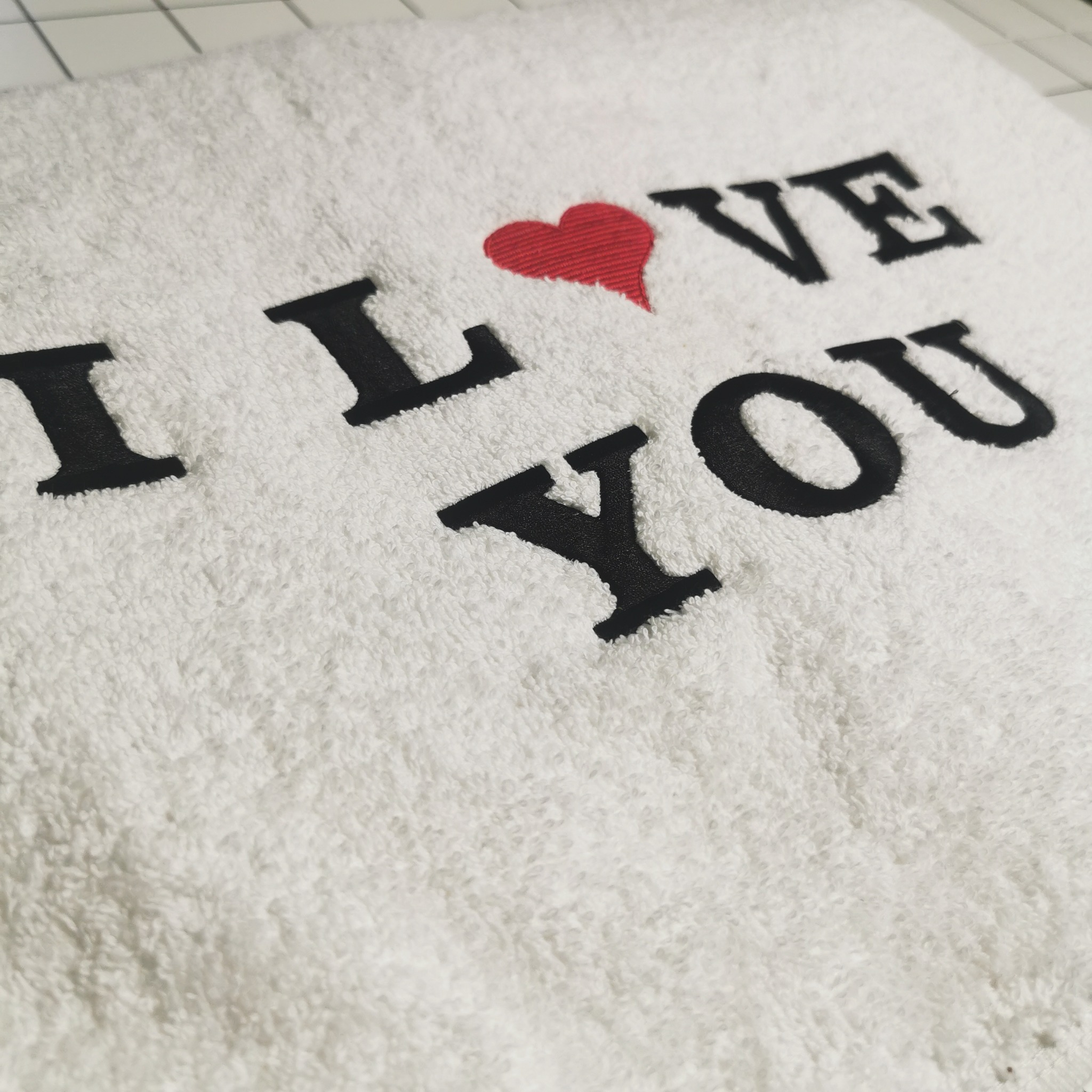 Biały ręcznik frotte z wyhaftowanym czarnym napisem 'I LOVE YOU' z czerwonym sercem zamiast 'LOVE', leżący na kafelkach. Detal haftu.