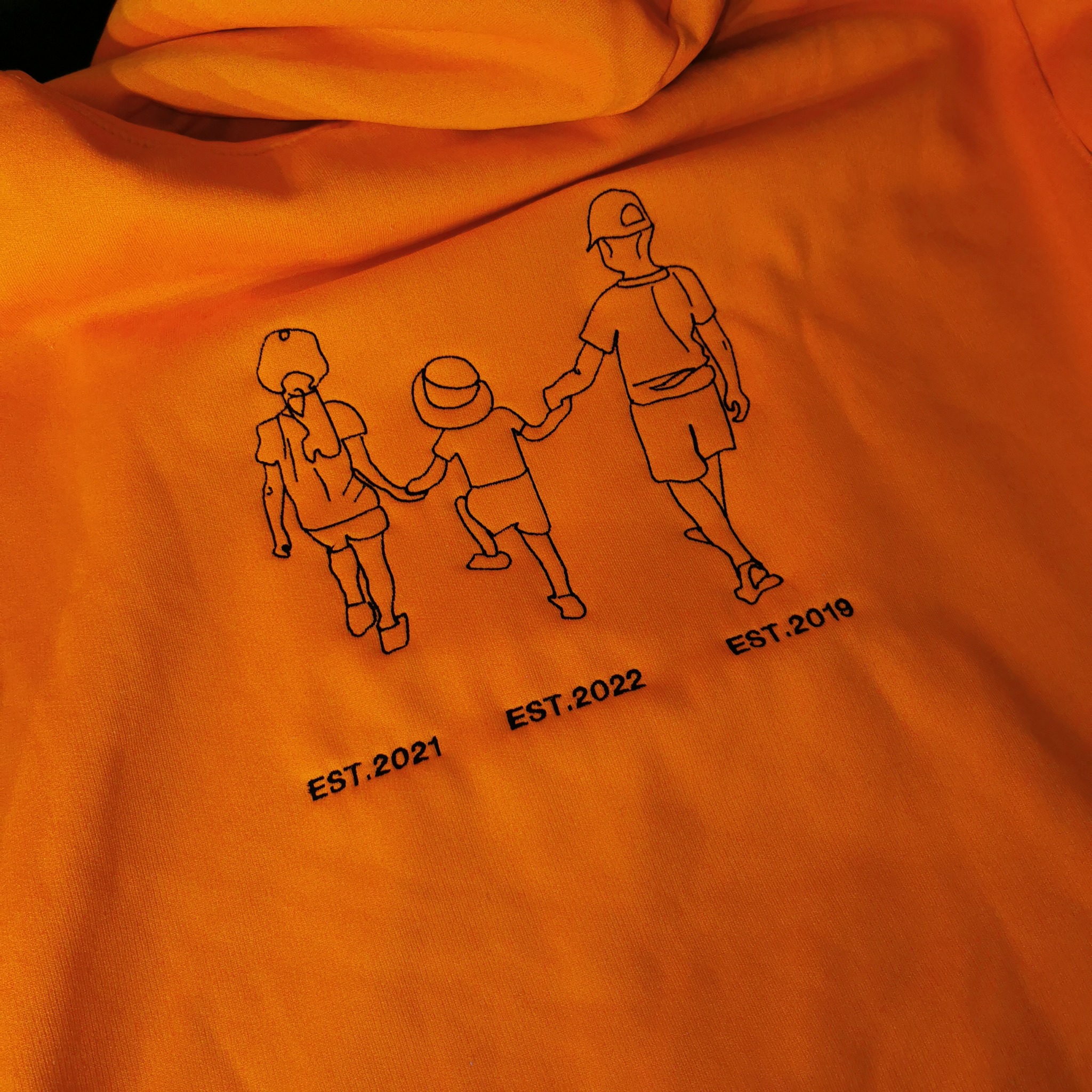Pomarańczowa bluza z kapturem, na której wyhaftowano czarną nicią rysunek trzech postaci i daty: EST.2021, EST.2022, EST.2019.