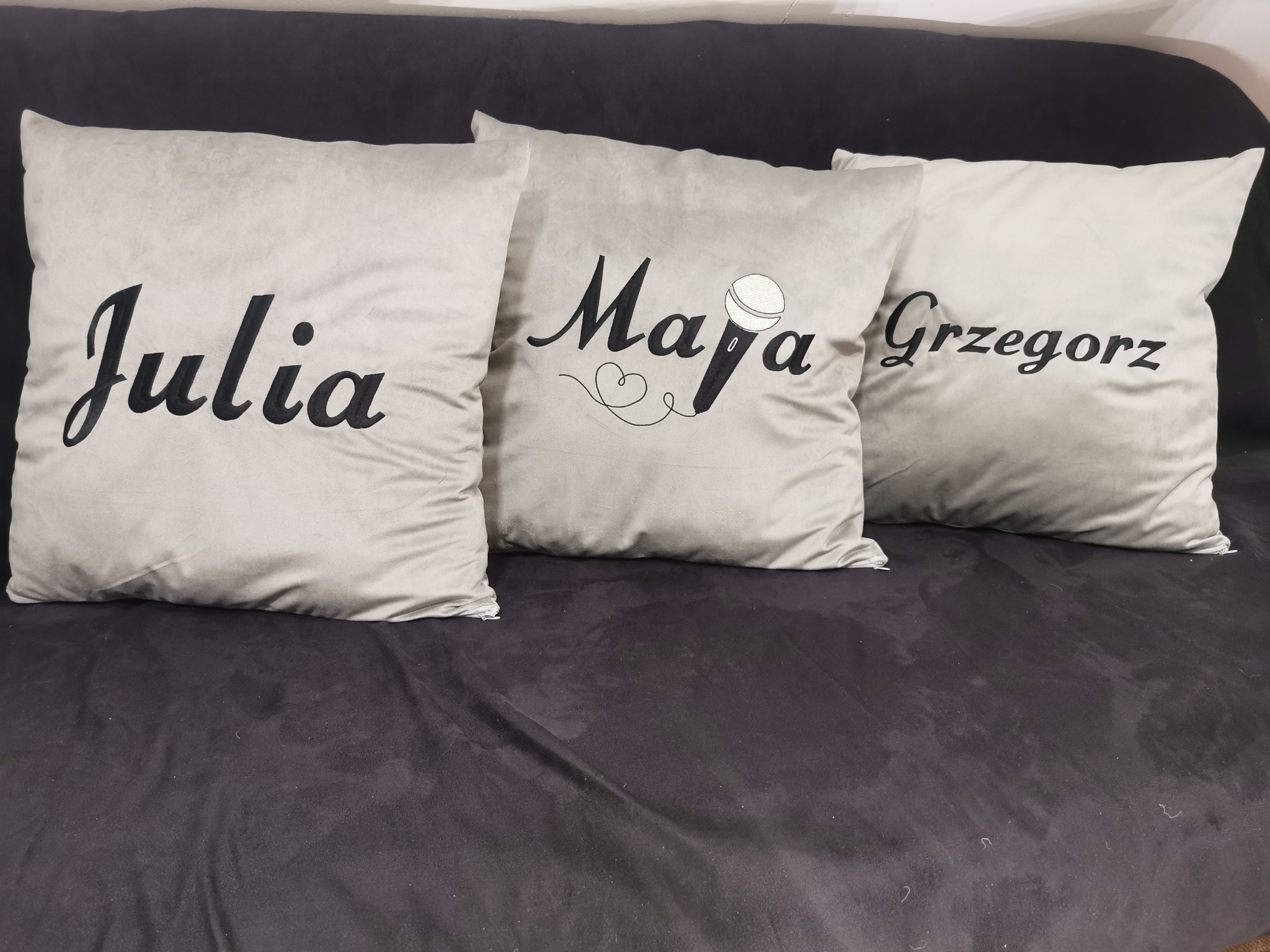 Dekoracyjne poduszki z imionami Julia, Maja (z mikrofonem) i Grzegorz, leżące na ciemnej kanapie. Imiona wykonane czarną czcionką, elegancki krój pisma.