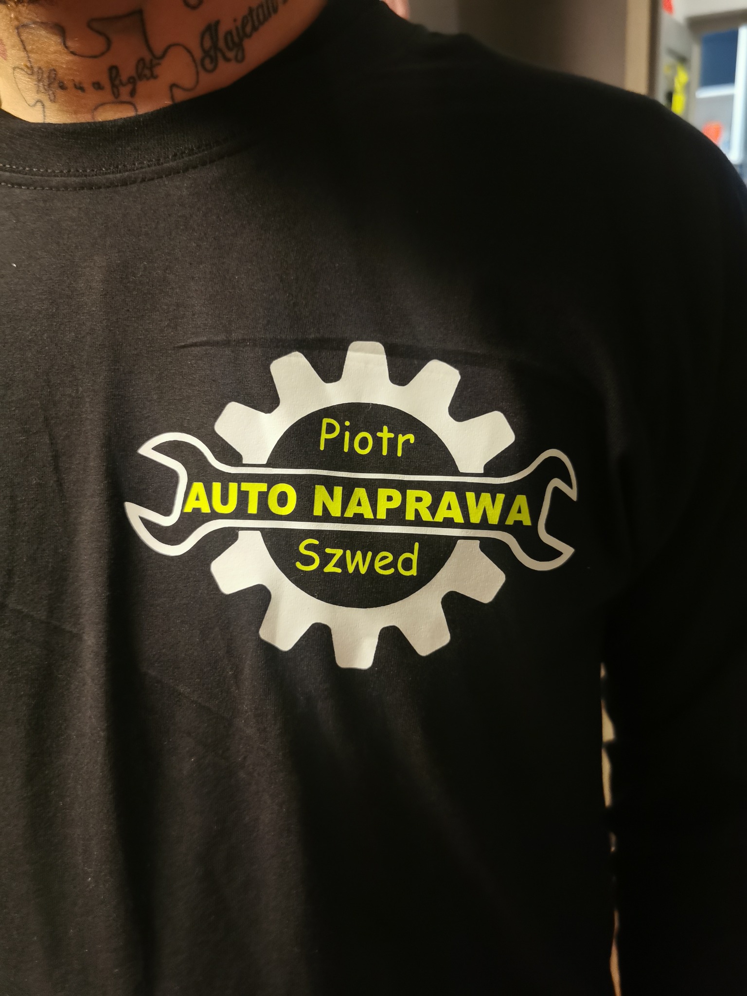 Czarna koszulka z nadrukiem logo warsztatu samochodowego, widoczny tatuaż na szyi modela. Żółto-biały nadruk z nazwą 'Auto Naprawa Piotr Szwed'.