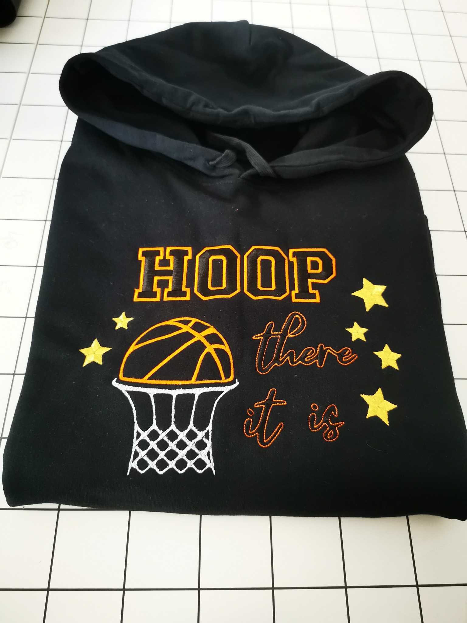 Czarna bluza z kapturem, zdobiona haftem z motywem koszykówki, napisem i gwiazdkami. Widoczne detale haftu i faktura materiału. Leży na białym tle w kratkę.