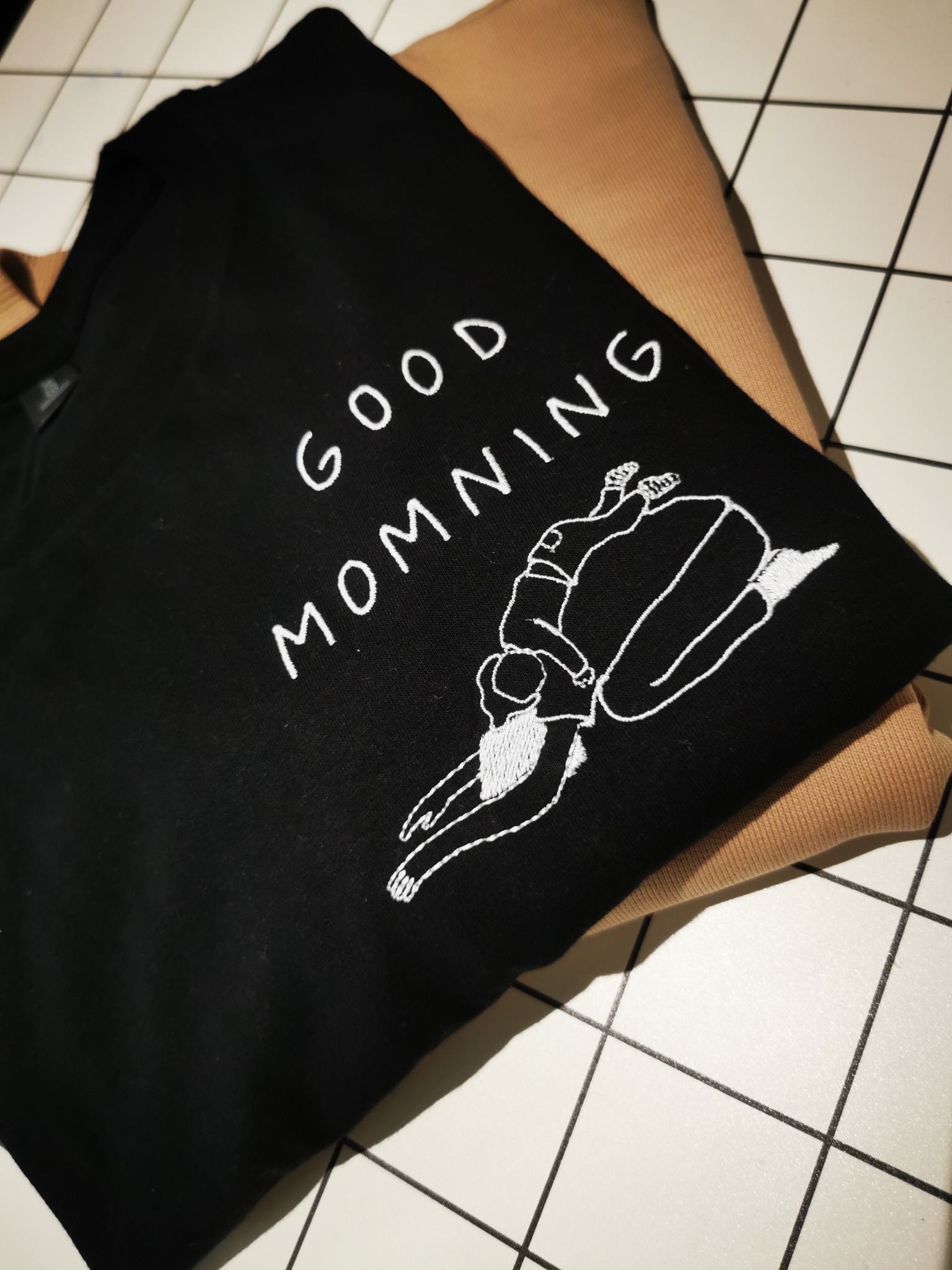 Czarna bluza z białym haftem przedstawiającym leżącą postać i napis 'GOOD MOMMING', ułożona na beżowej bluzie na tle białych płytek.