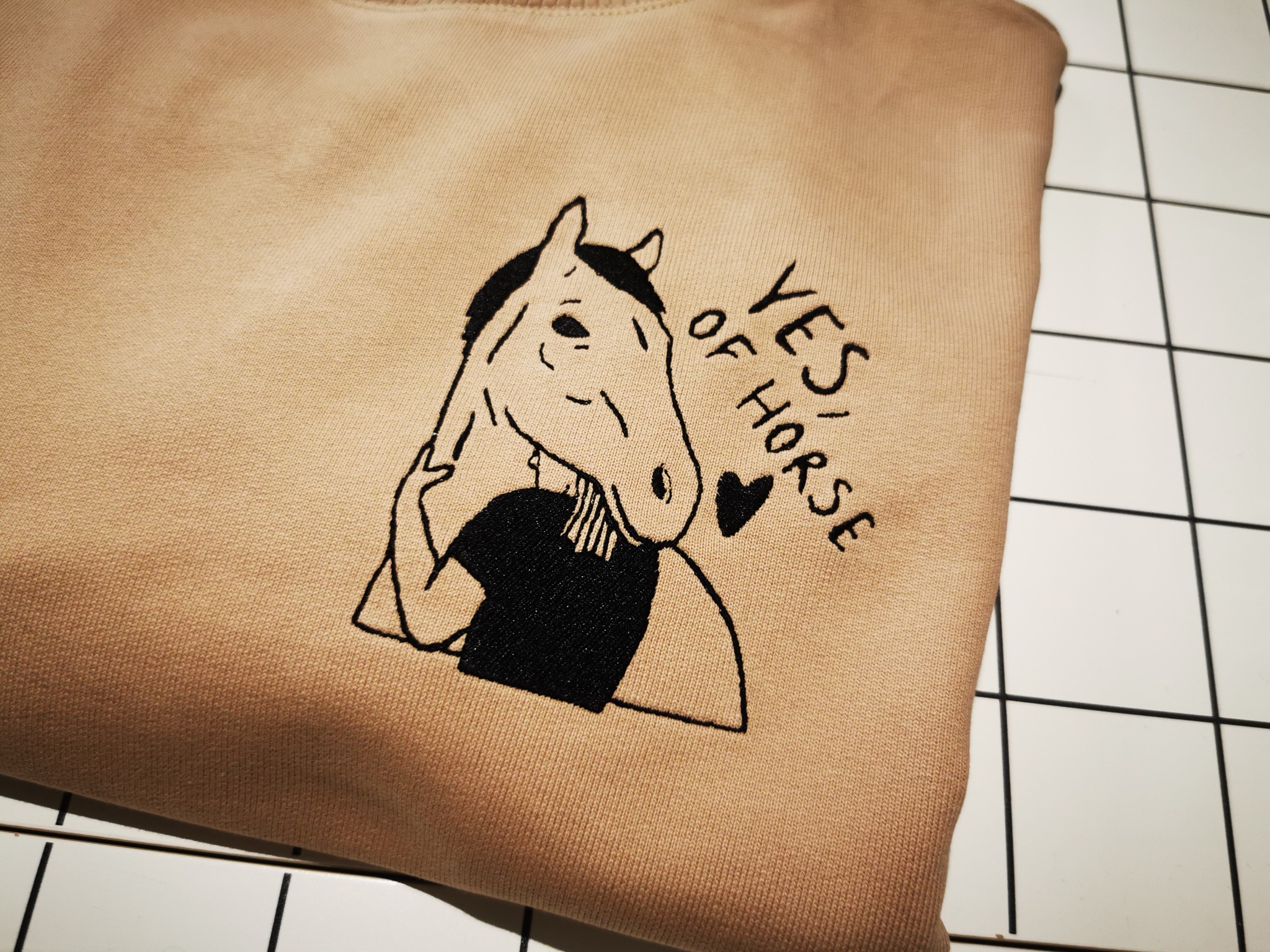 Beżowa bluza z haftem przedstawiającym konia w koszuli i napisem 'Yes of Horse' leży na białej, kafelkowej powierzchni.