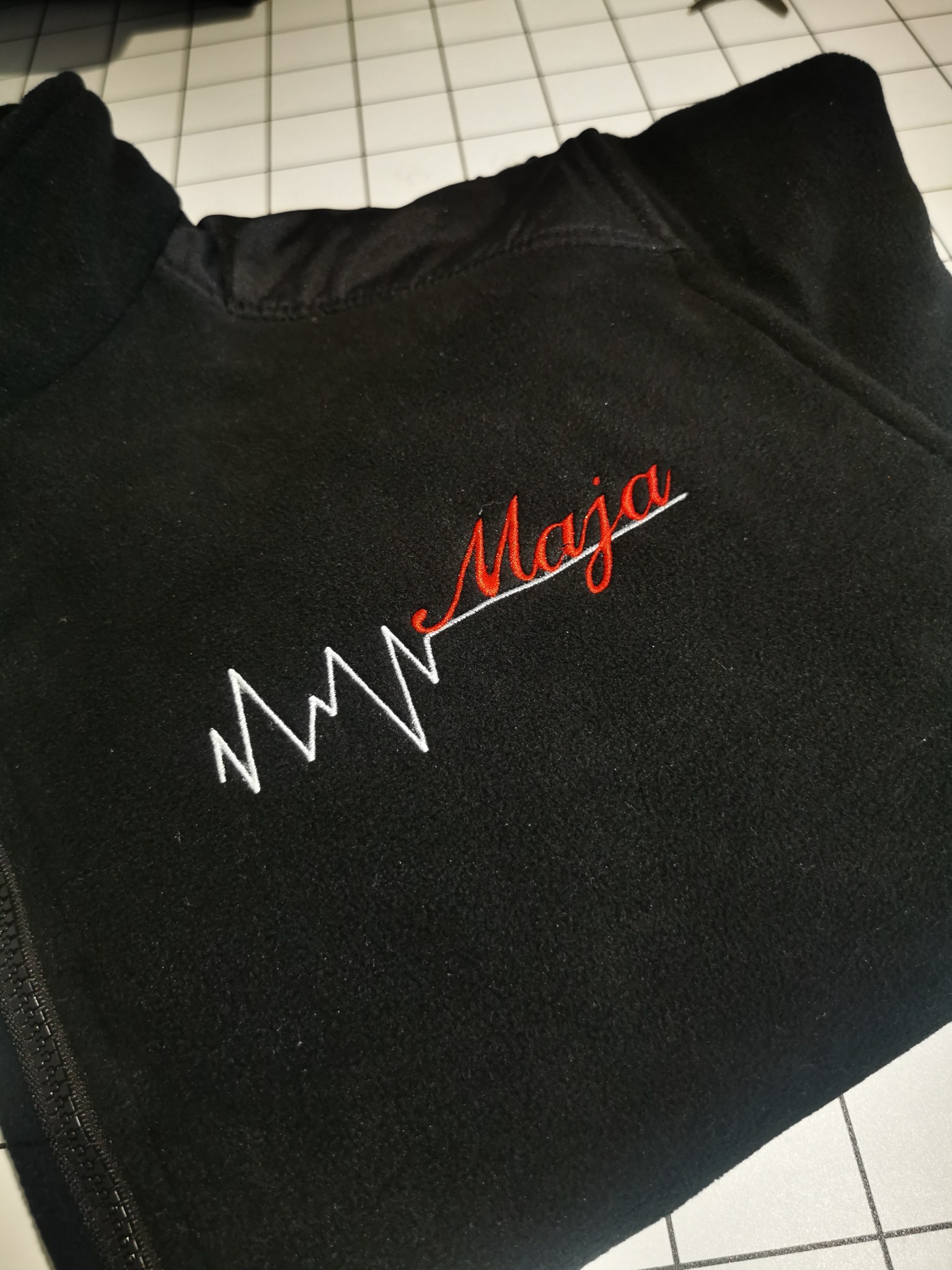 Czarna bluza polarowa z haftowanym, czerwonym napisem 'Maja' i białym motywem EKG. Detal haftu na tle kratkowanej powierzchni.