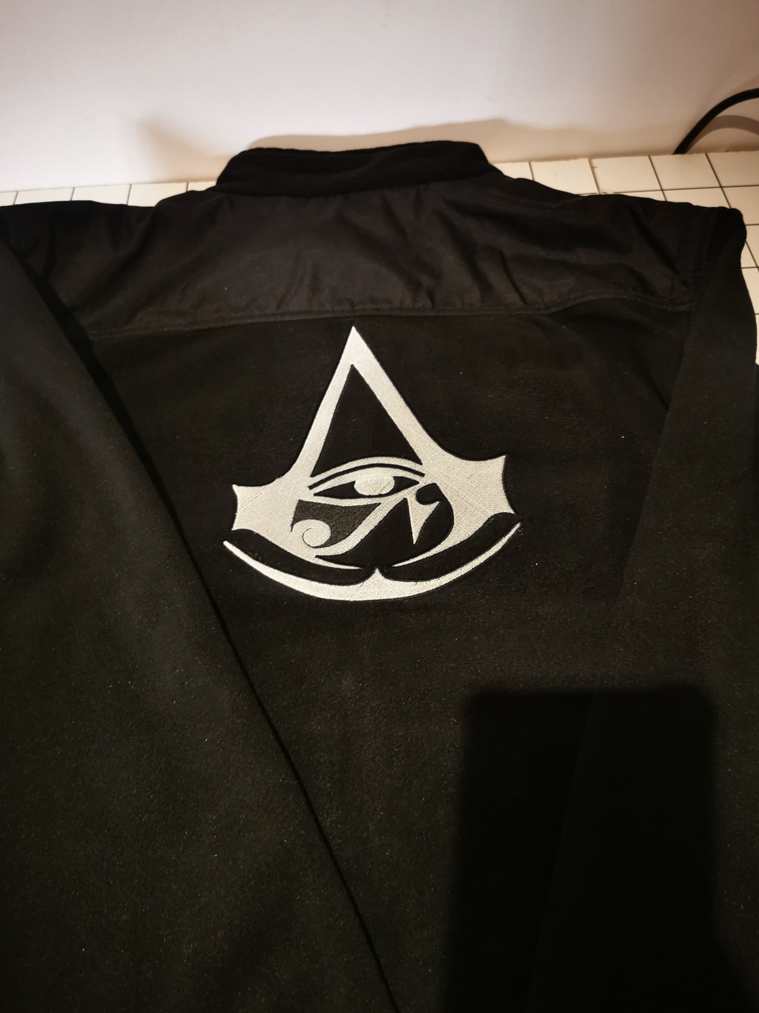 Czarna kurtka z dużym, srebrnym haftowanym logo inspirowanym grą Assassin's Creed na plecach. Widok z tyłu, ukazujący detale haftu i materiału.