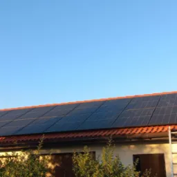 Panele fotowoltaiczne na dachu budynku z czerwoną dachówką, widoczne z dołu, z drabiny opartej o rynnę i zielenią na pierwszym planie, w słoneczny dzień.