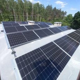 Inhelio sp. z o. o. - Panel fotowoltaiczny na dachu budynku, widok z góry, w tle drzewa. Układ paneli w rzędach, szara płaska powierzchnia dachu, kominy wentylacyjne.