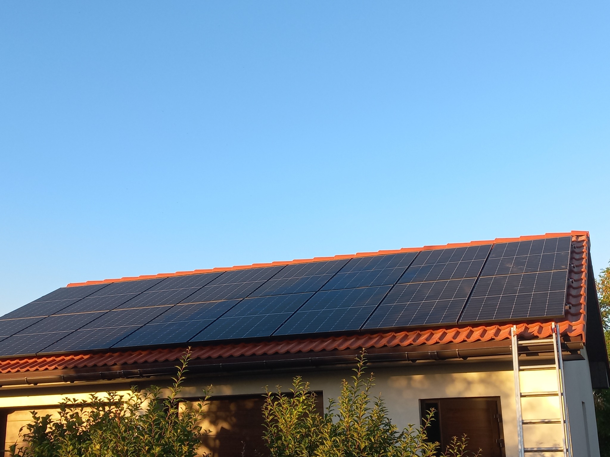 Panele fotowoltaiczne na dachu budynku z czerwoną dachówką, widoczne z dołu, z drabiny opartej o rynnę i zielenią na pierwszym planie, w słoneczny dzień.