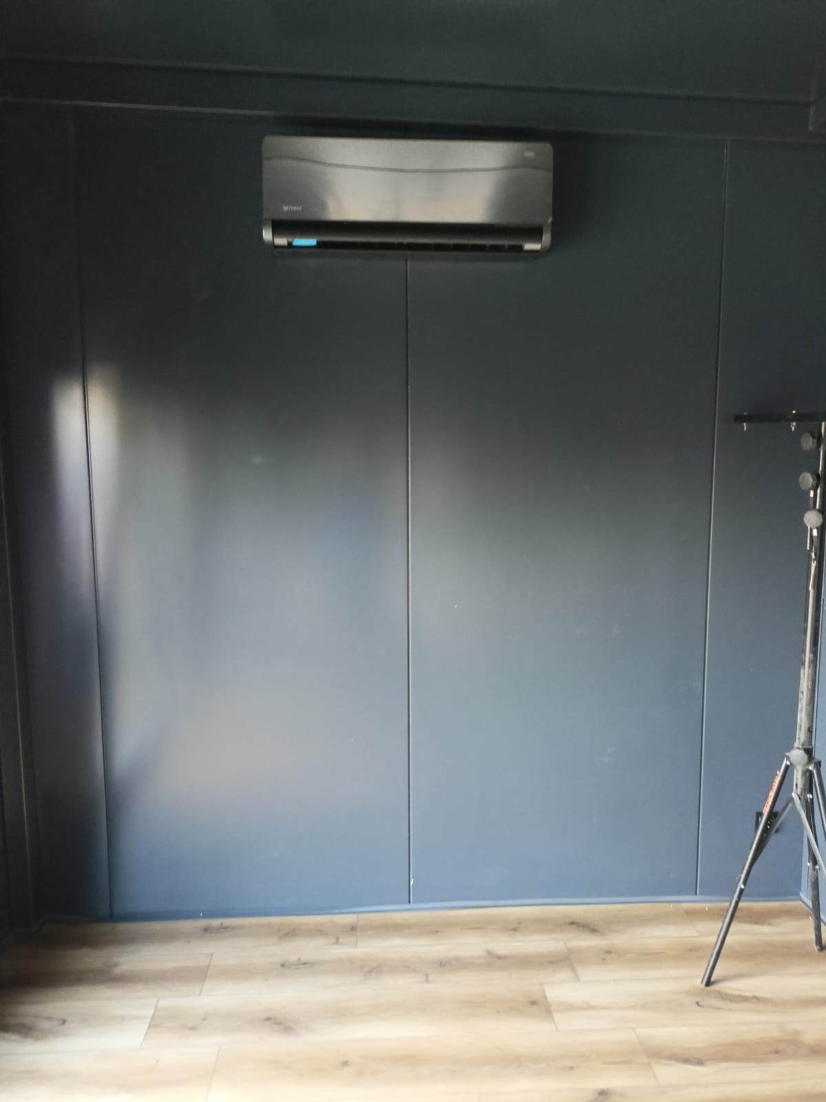 Zamontowany klimatyzator na ciemnej ścianie z panelami, widoczna podłoga z paneli i fragment statywu oświetleniowego po prawej stronie.